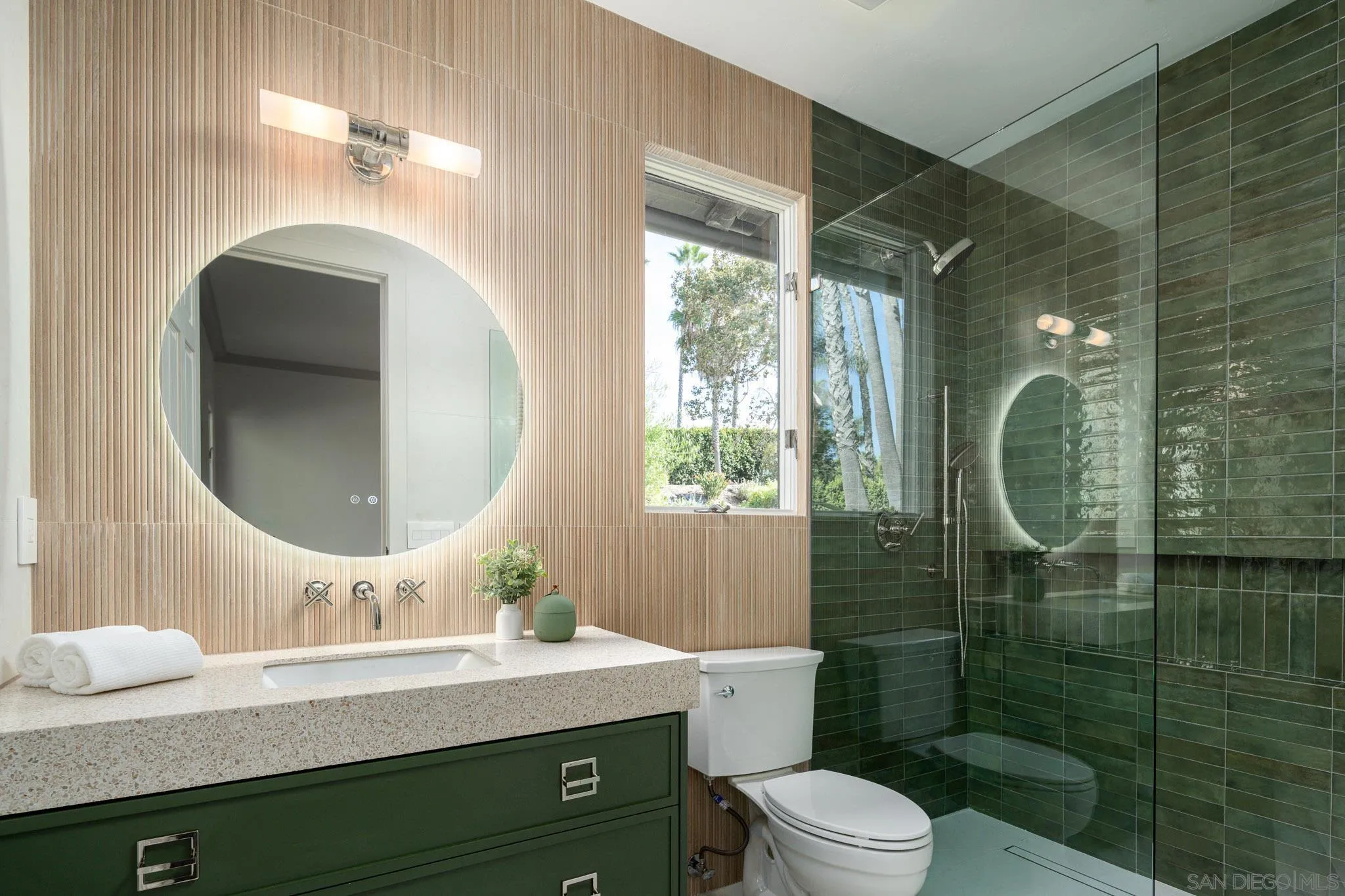 Bathroom with a green-tiled glass shower, dark green vanity, and round mirror beside a window.