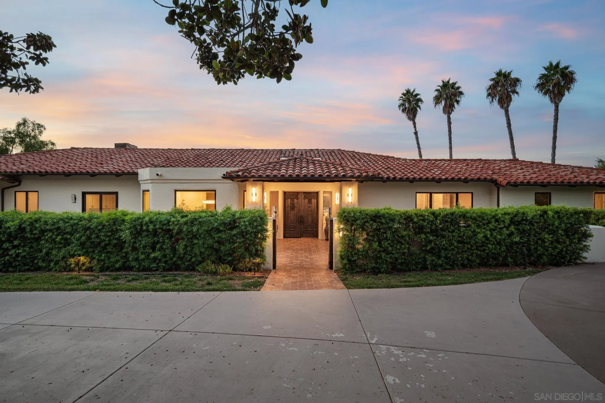 Single-story stucco home with a red tile roof and warmly lit, hedge-framed entrance at dusk.