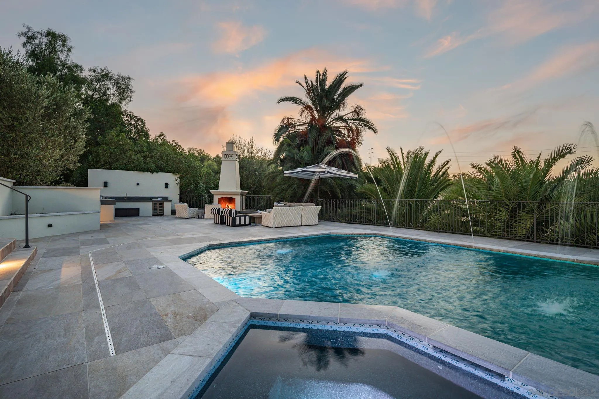 Outdoor pool and raised spa beside a patio fireplace, lounge seating, and palm trees at dusk.