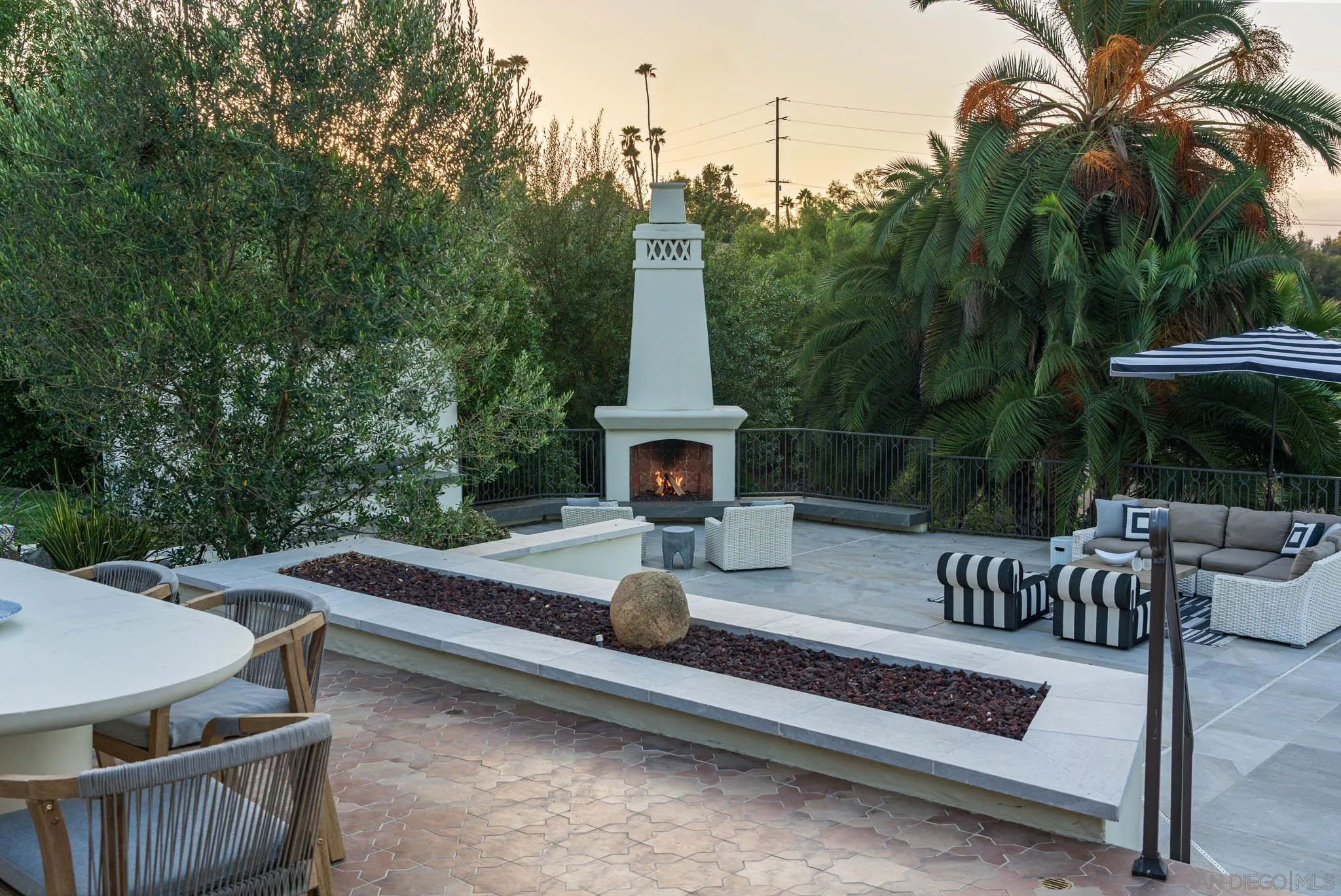 Outdoor patio with a lit fireplace, dining table, and lounge seating surrounded by palm trees.