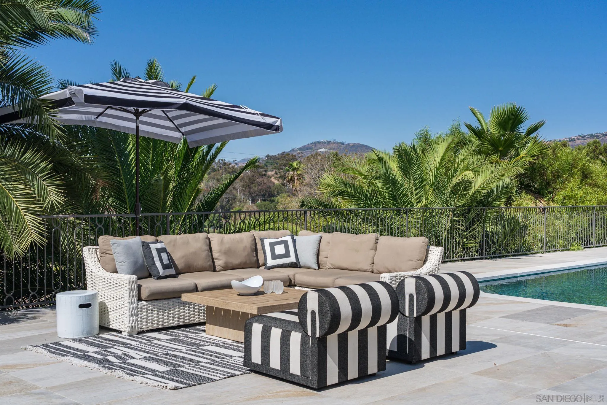 Poolside patio with wicker seating, striped ottomans, and a hillside view.