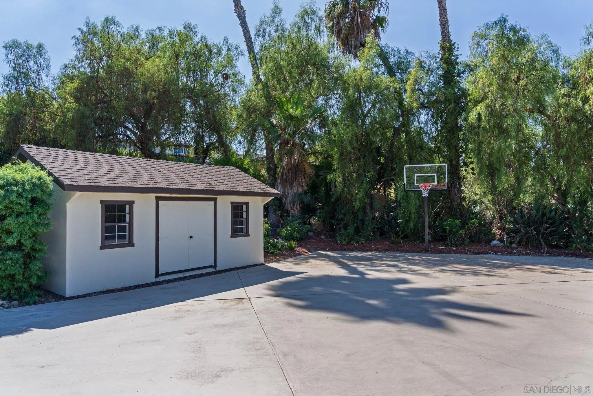 Concrete basketball court beside a small white outbuilding, with a hoop and mature trees.
