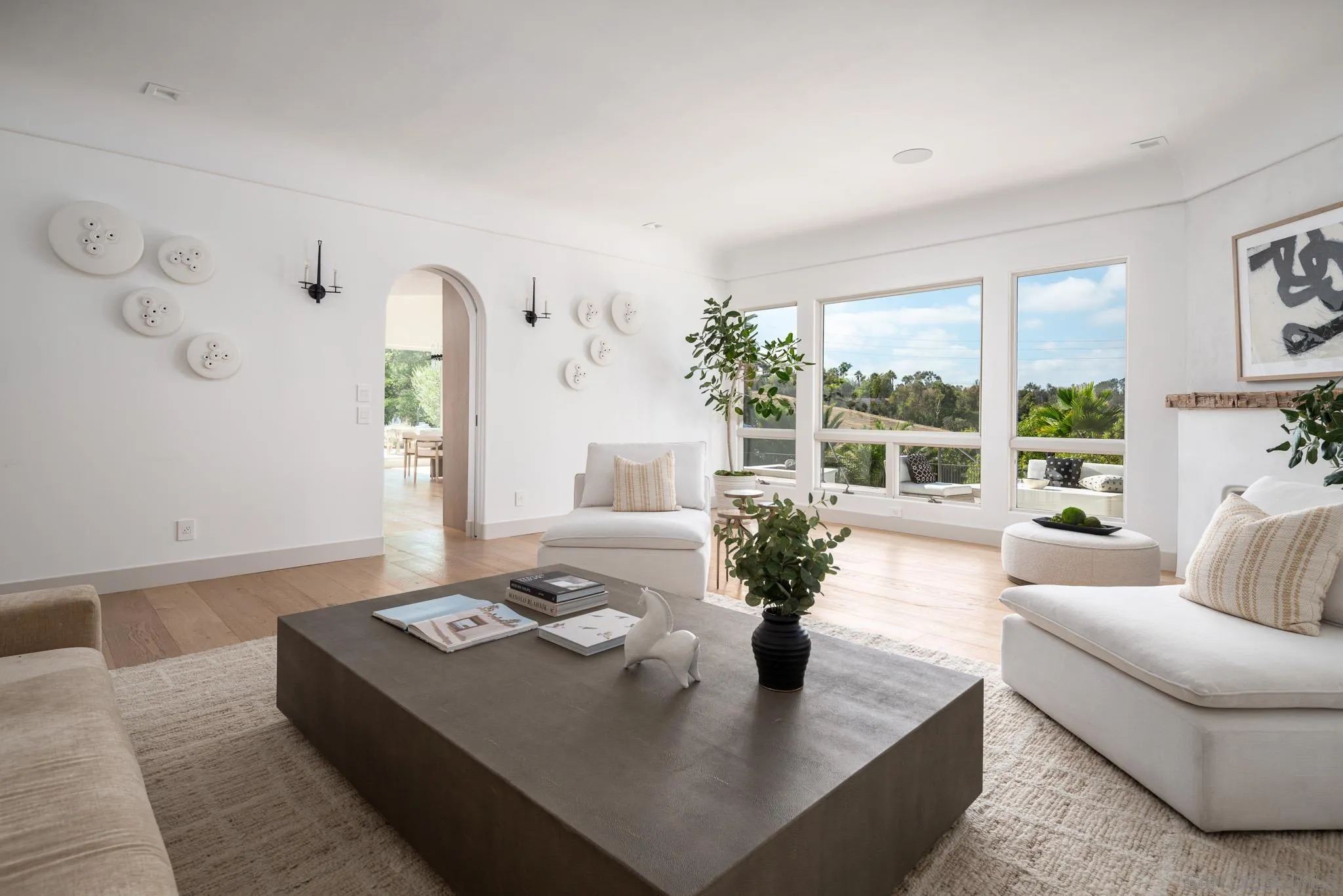 Bright living room with white sofas, a large dark coffee table, and wide windows overlooking greenery.