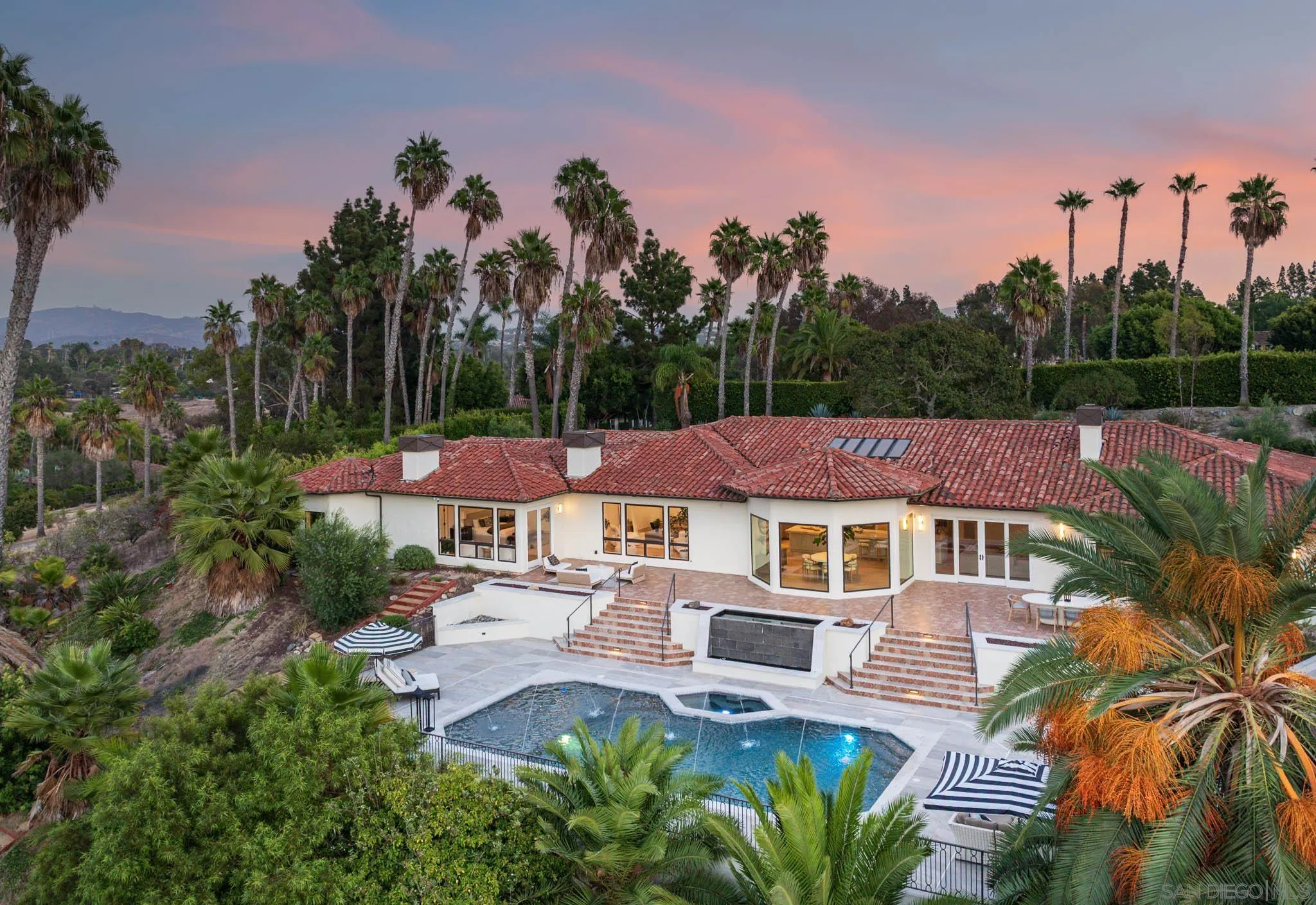 Home and swimming pool surrounded by palm trees, pictured at dusk with distant hills.
