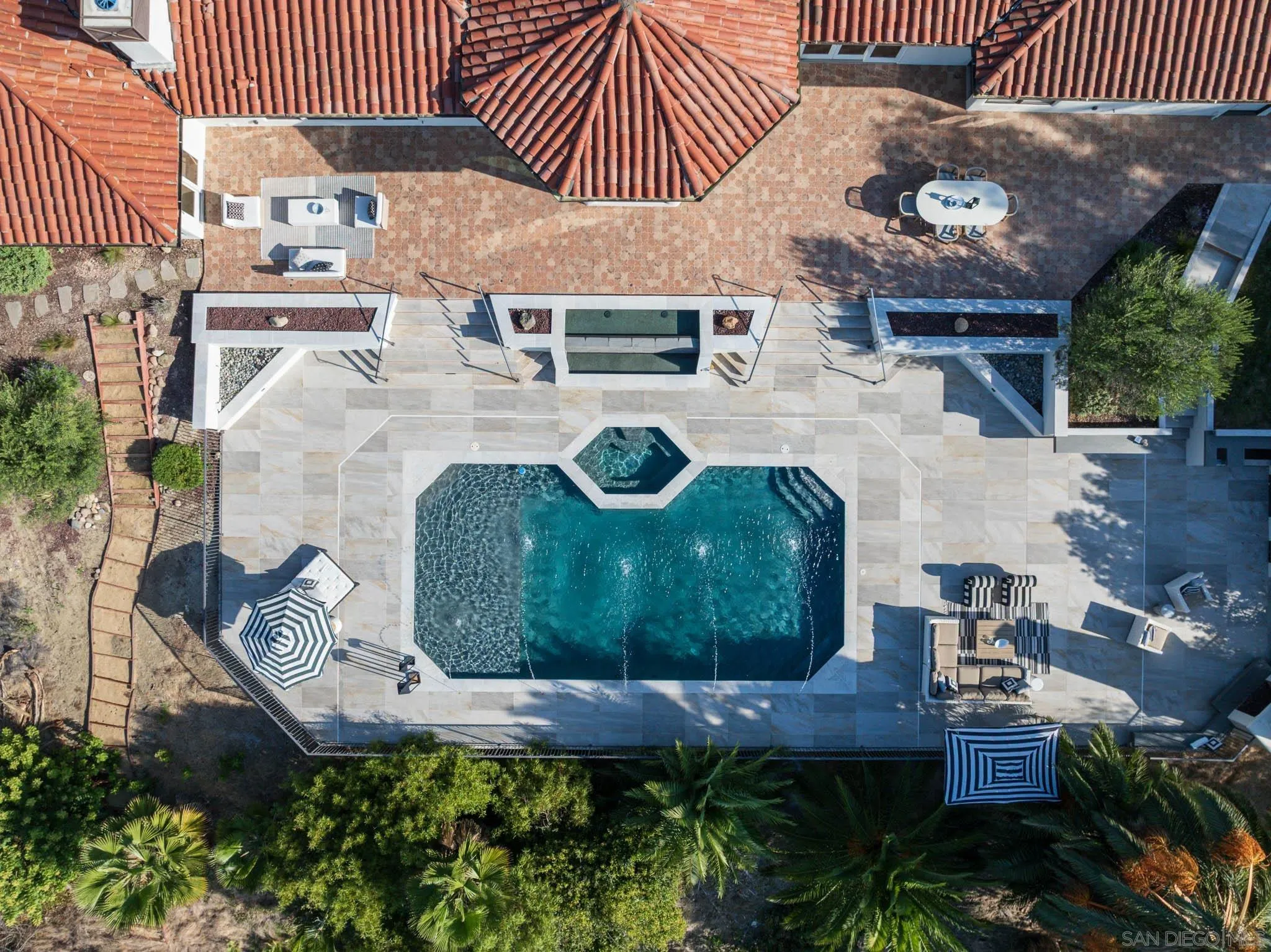 Aerial view of a geometric backyard pool with an attached spa, pale stone patio, and striped umbrellas.