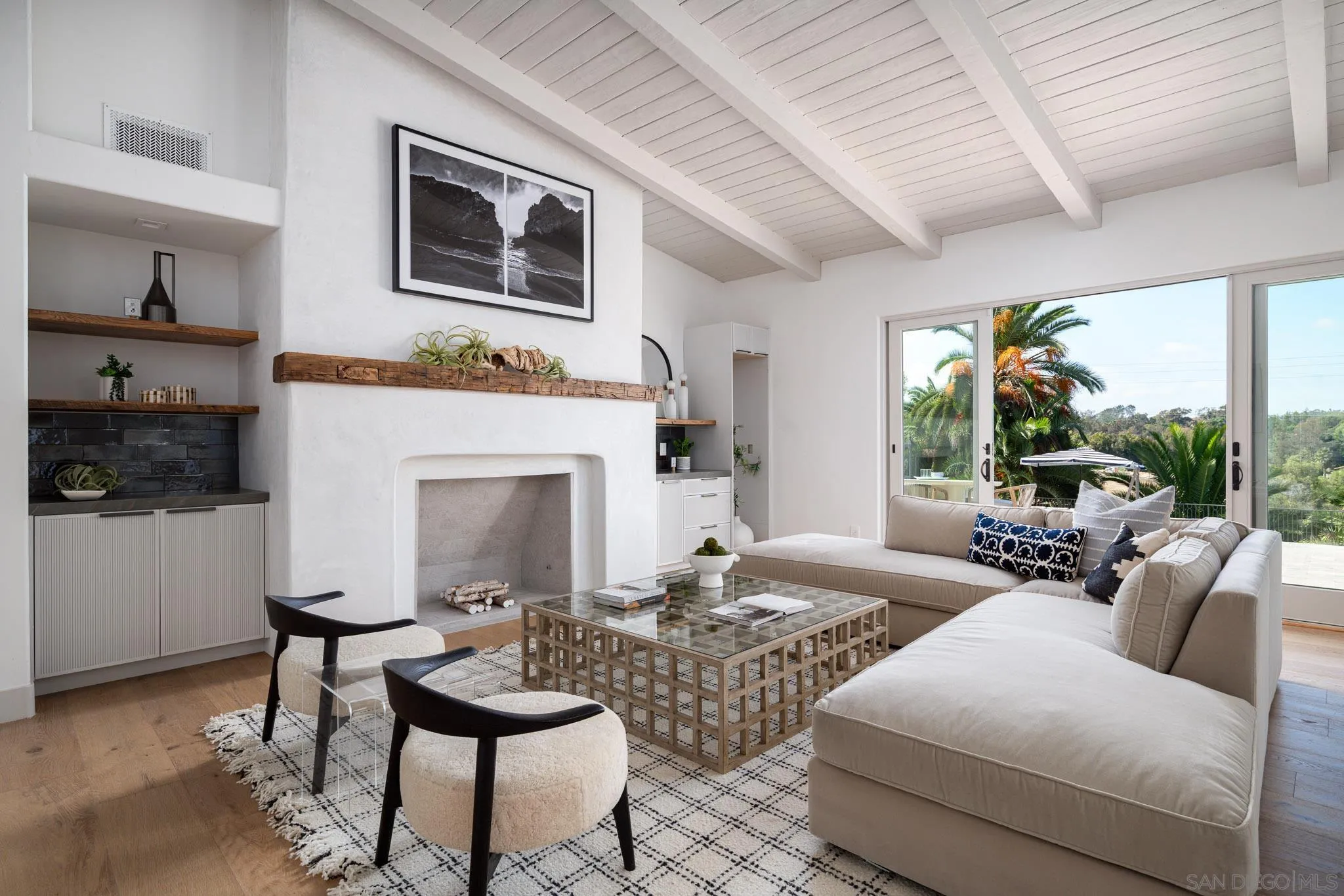 Staged living room with a vaulted beamed ceiling, white fireplace, and sliding doors overlooking palms.