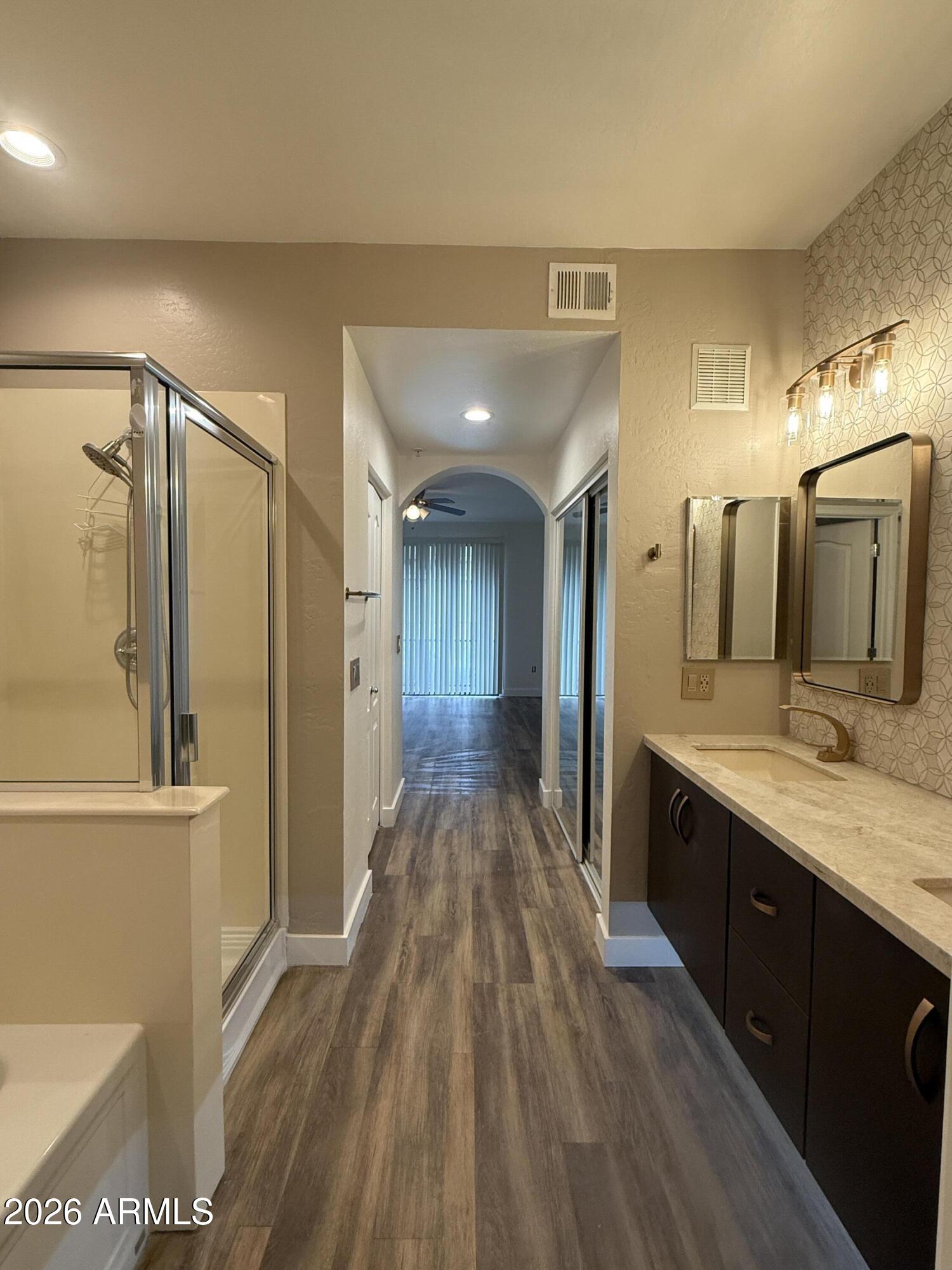 Bathroom with glass shower, long dark vanity, and wood-look flooring leading into a hallway.