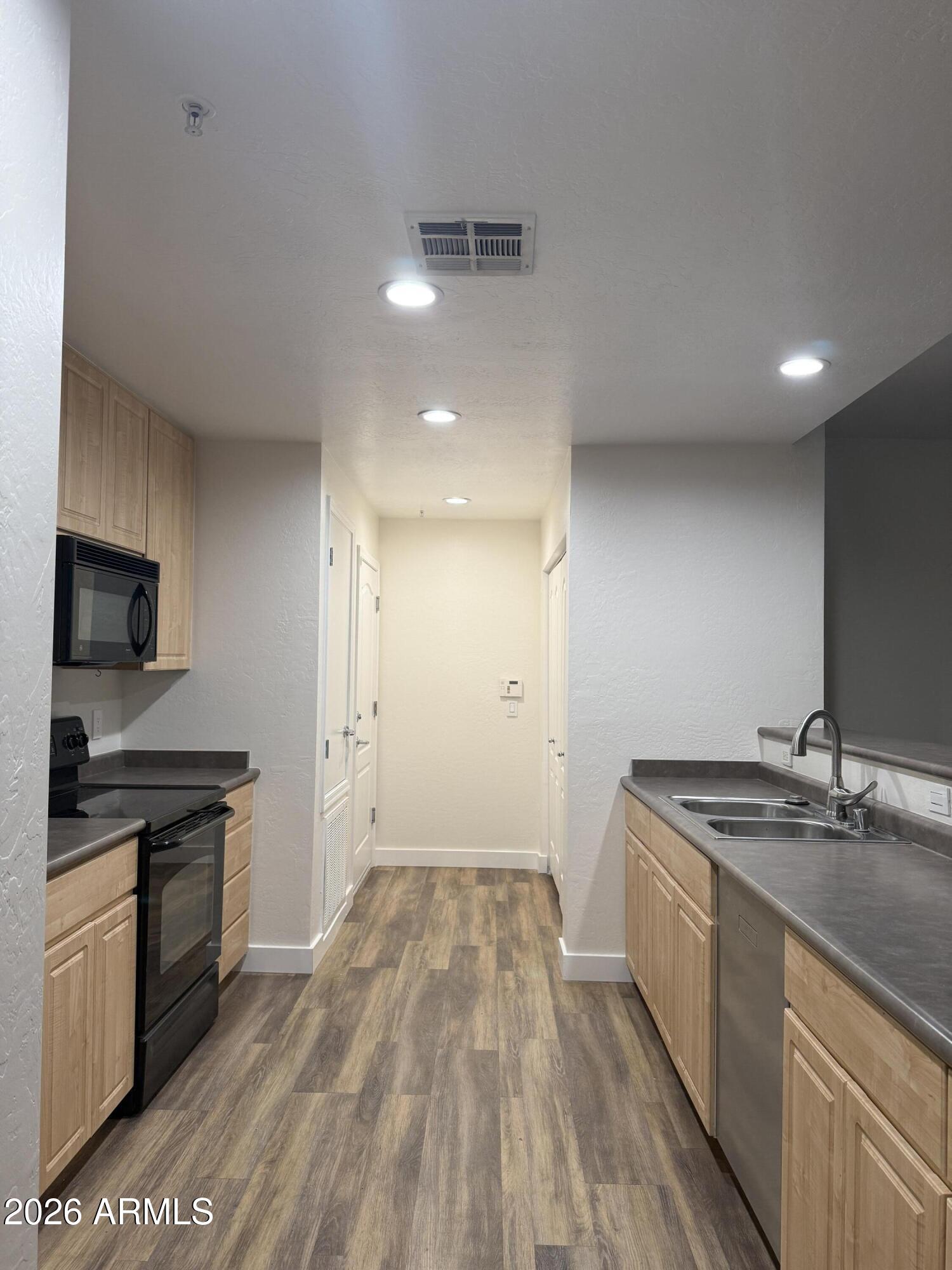 Galley kitchen with light wood cabinets, dark counters, black appliances, and a double-basin sink.