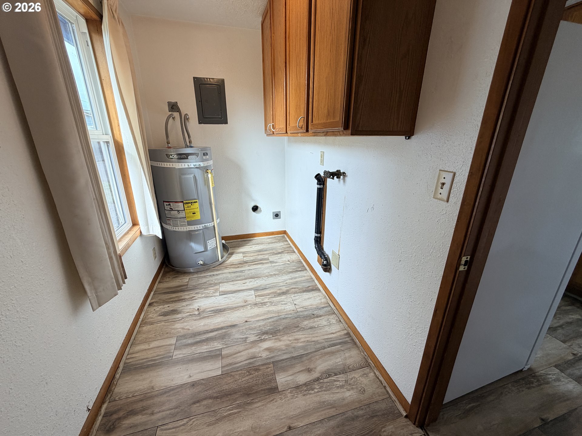Utility room with a water heater, exposed plumbing, and overhead wooden cabinets.
