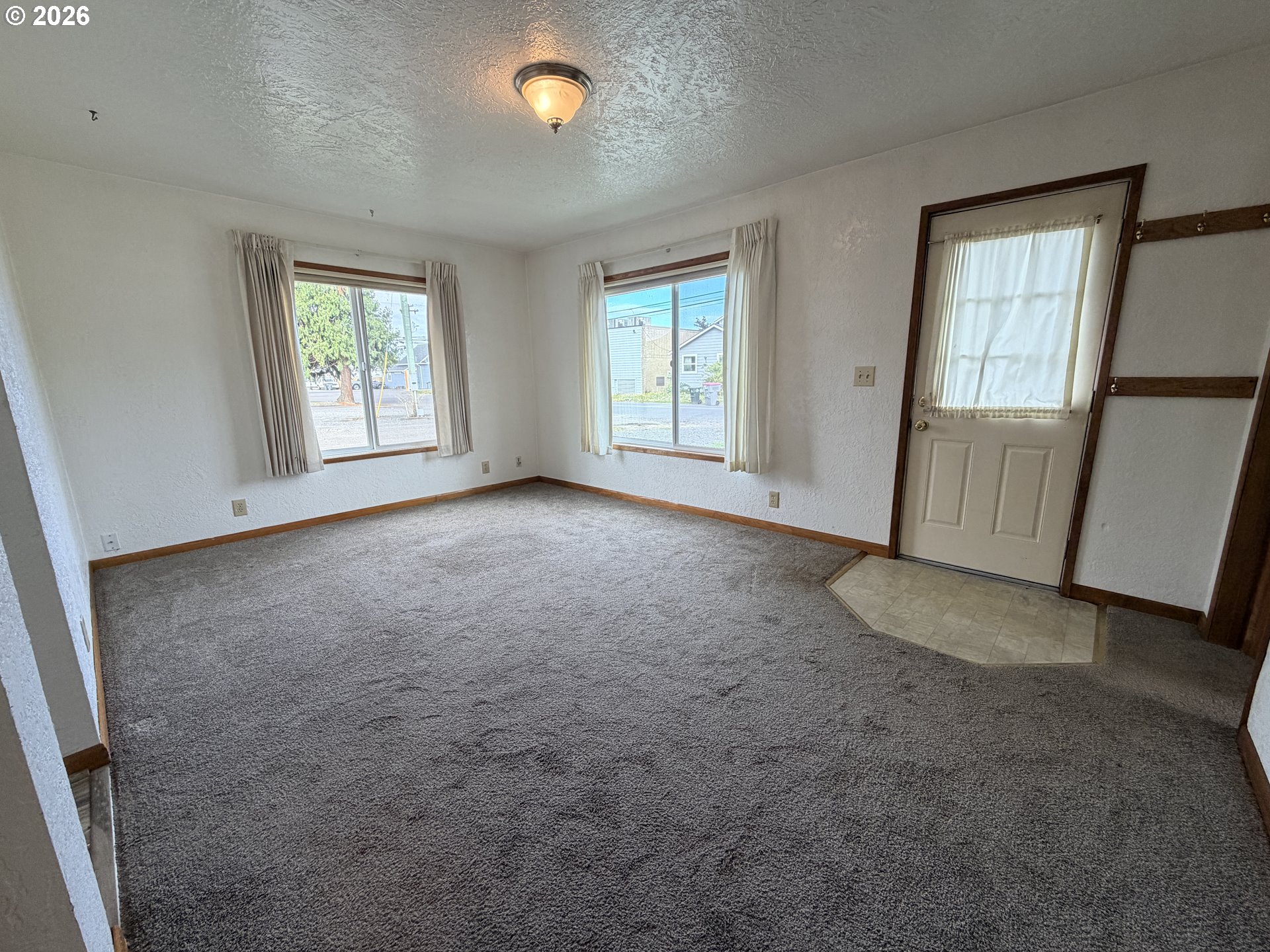 Empty carpeted living room with two curtained windows and an exterior door.