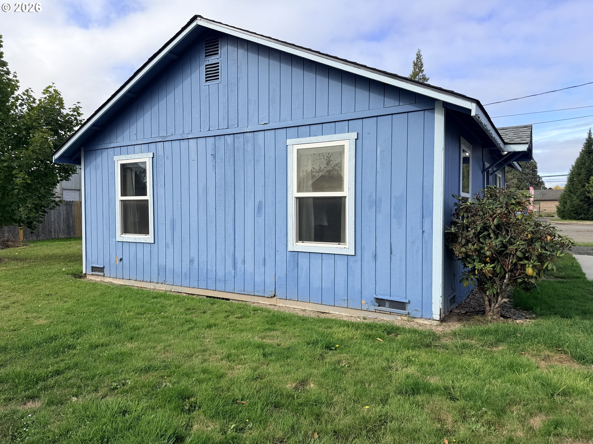 Blue-sided building with white-trimmed windows beside a grassy lawn and shrubs.
