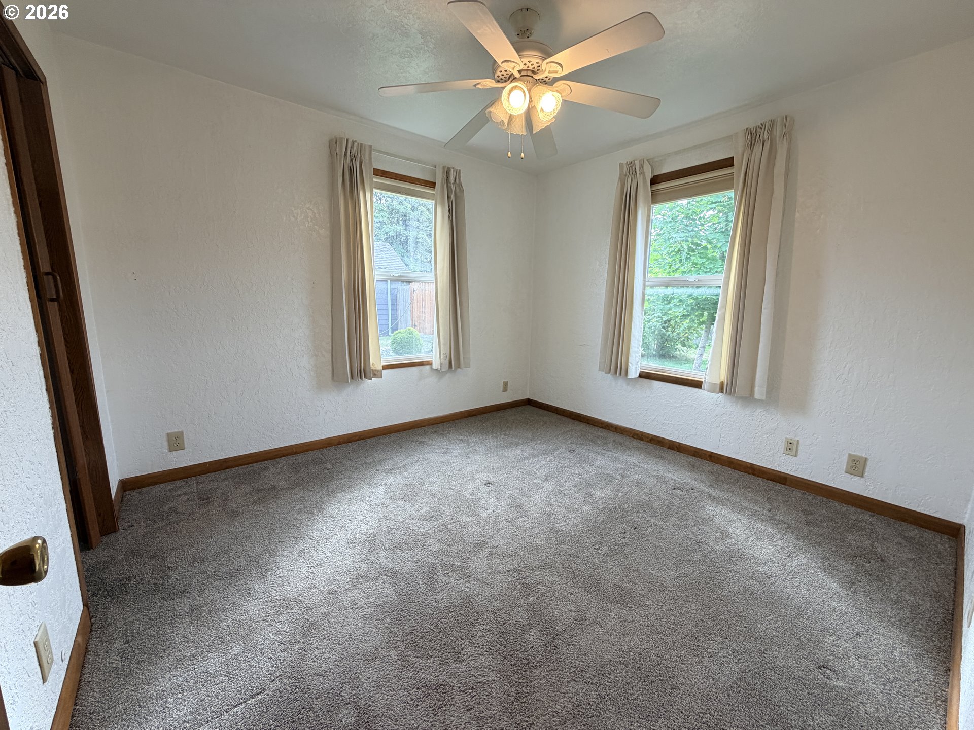 Empty carpeted room with two curtained windows and a ceiling fan with lights.