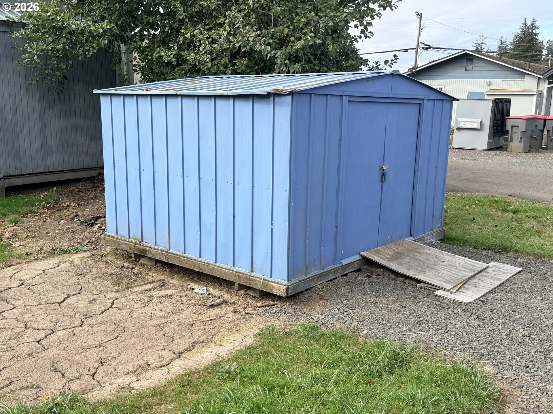Blue metal storage shed with double doors and plywood sheets forming a ramp at the entrance.