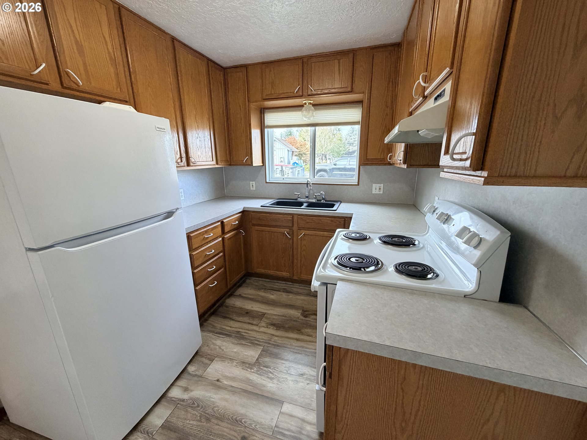 Compact kitchen with wood cabinets, white appliances, a window over the sink, and wood-look flooring.