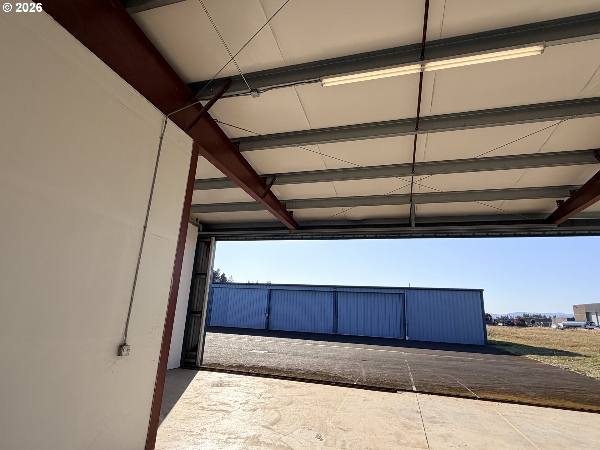 Airport hangar interior with an open door, exposed steel beams, and a blue hangar across the apron.