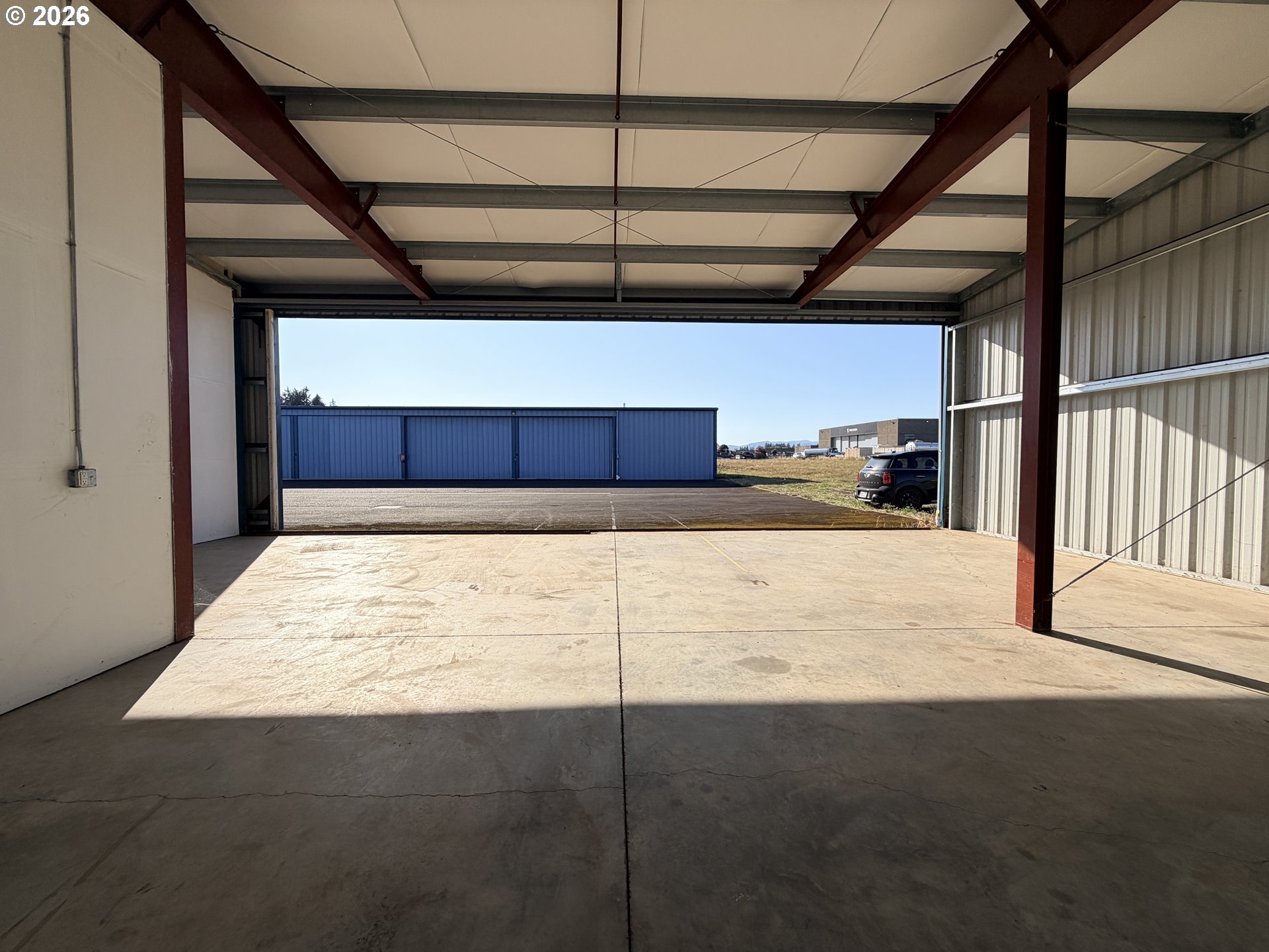 Empty airport hangar with exposed steel beams and a wide-open door facing blue hangars.