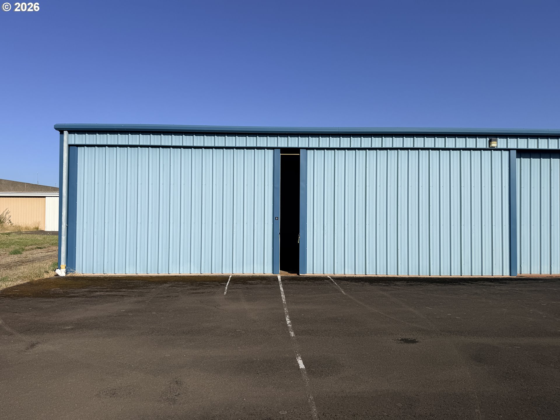Light-blue metal airport hangar with an open doorway and marked asphalt parking spaces.