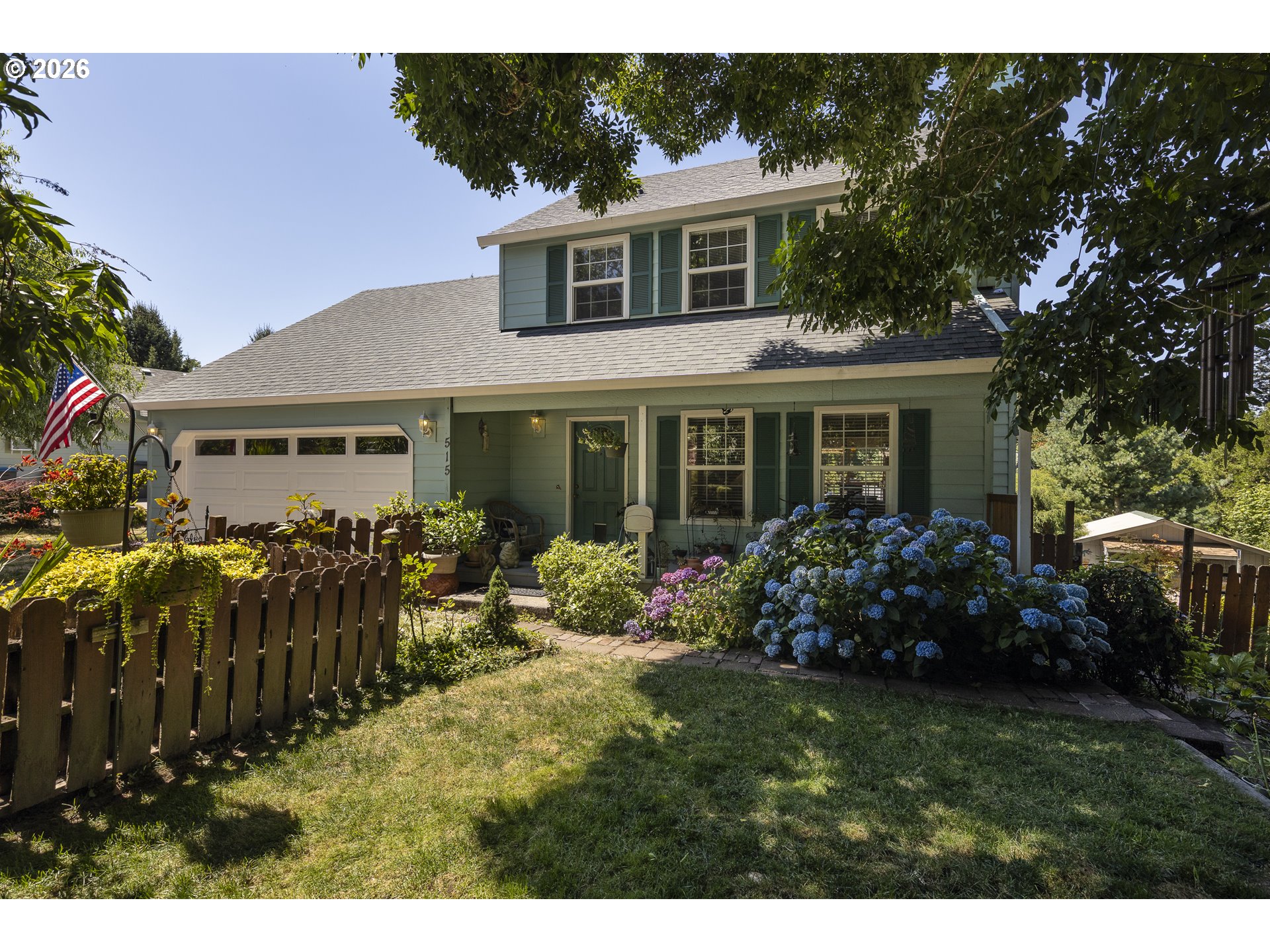Sage-green two-story house with a white garage, fenced yard, and blooming hydrangeas.