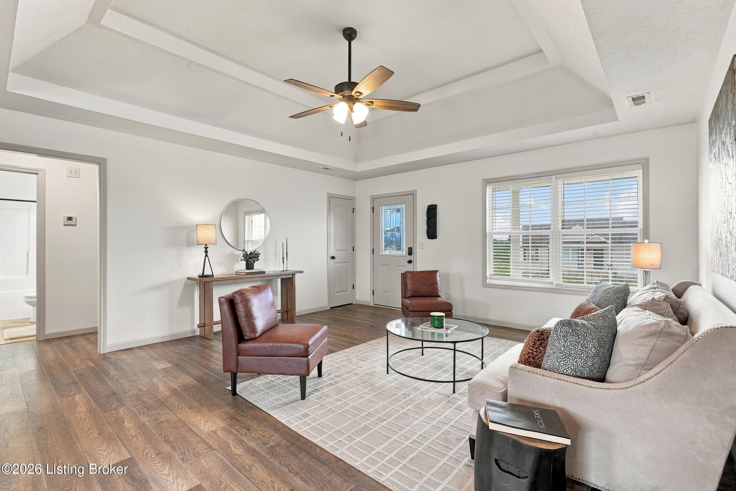 Staged living room with tray ceiling, brown leather chairs, light sofa, and large front window.