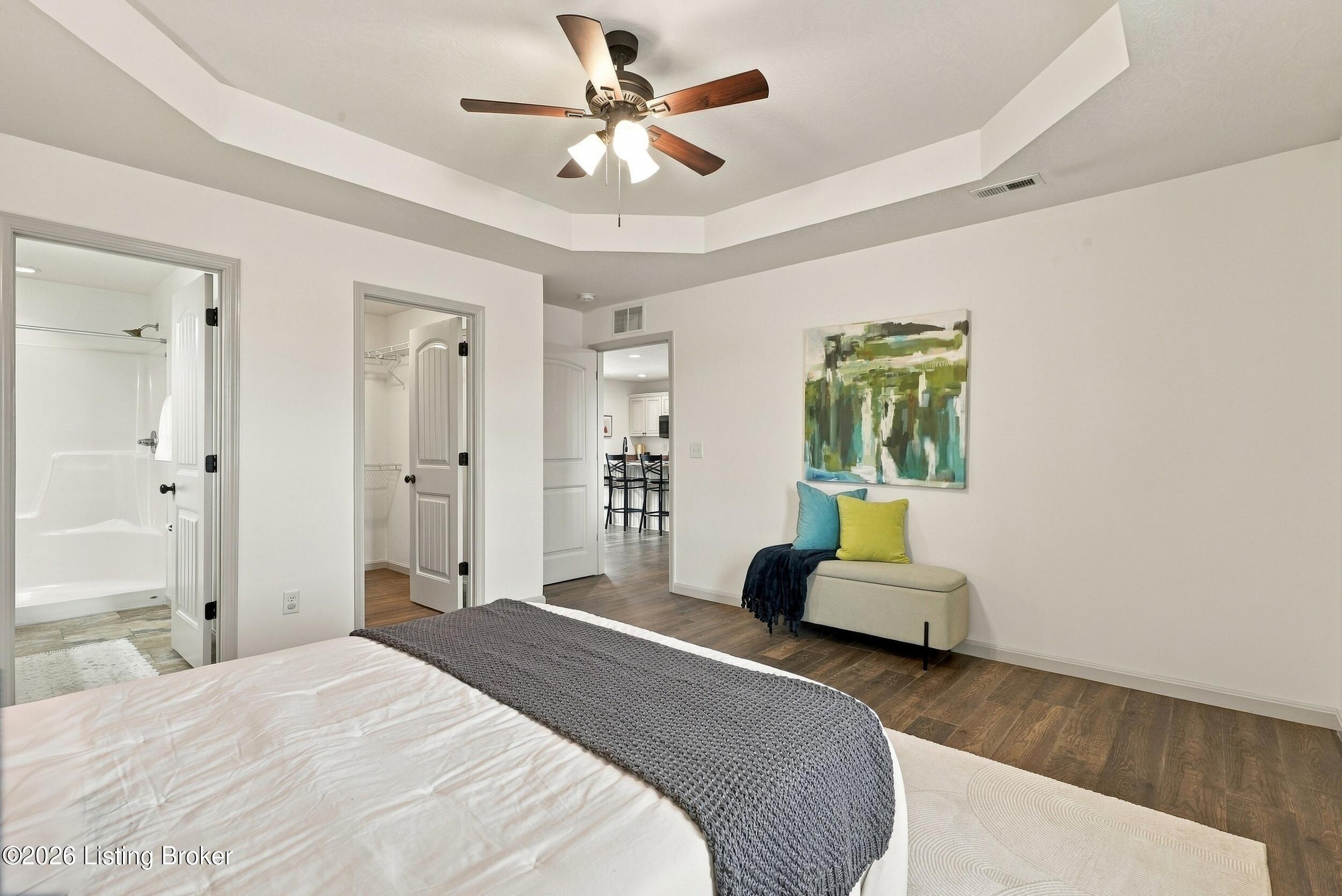 Furnished bedroom with tray ceiling, gray bedspread, colorful bench pillows, and open bathroom doorway.