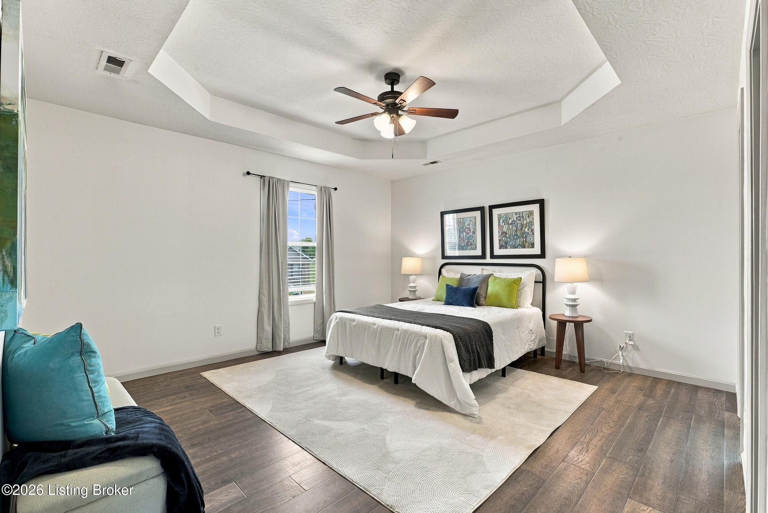 Staged bedroom with a white bed, colorful pillows, and a tray ceiling with a ceiling fan.