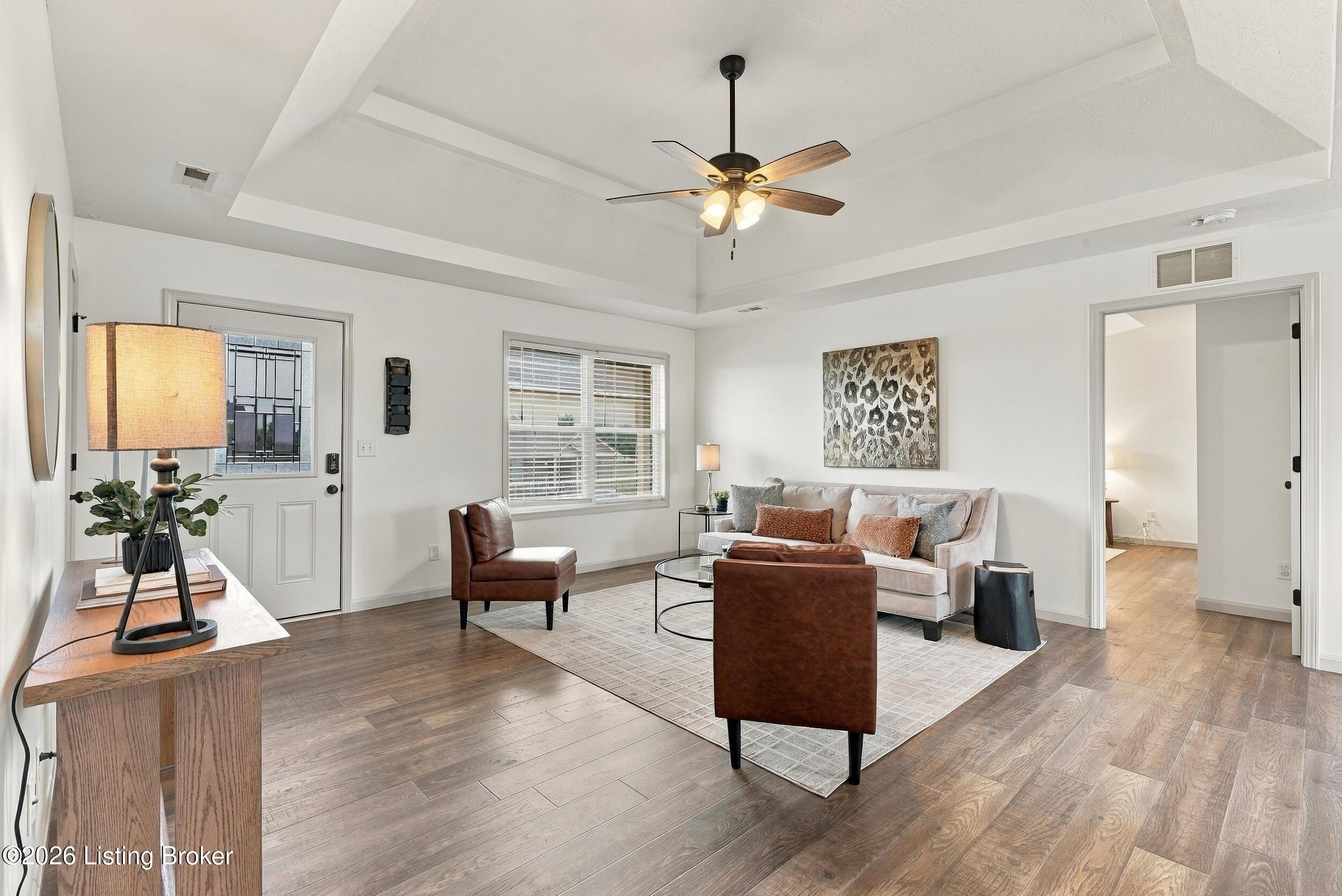 Staged living room with a tray ceiling, ceiling fan, wood-look flooring, and cream and brown seating.