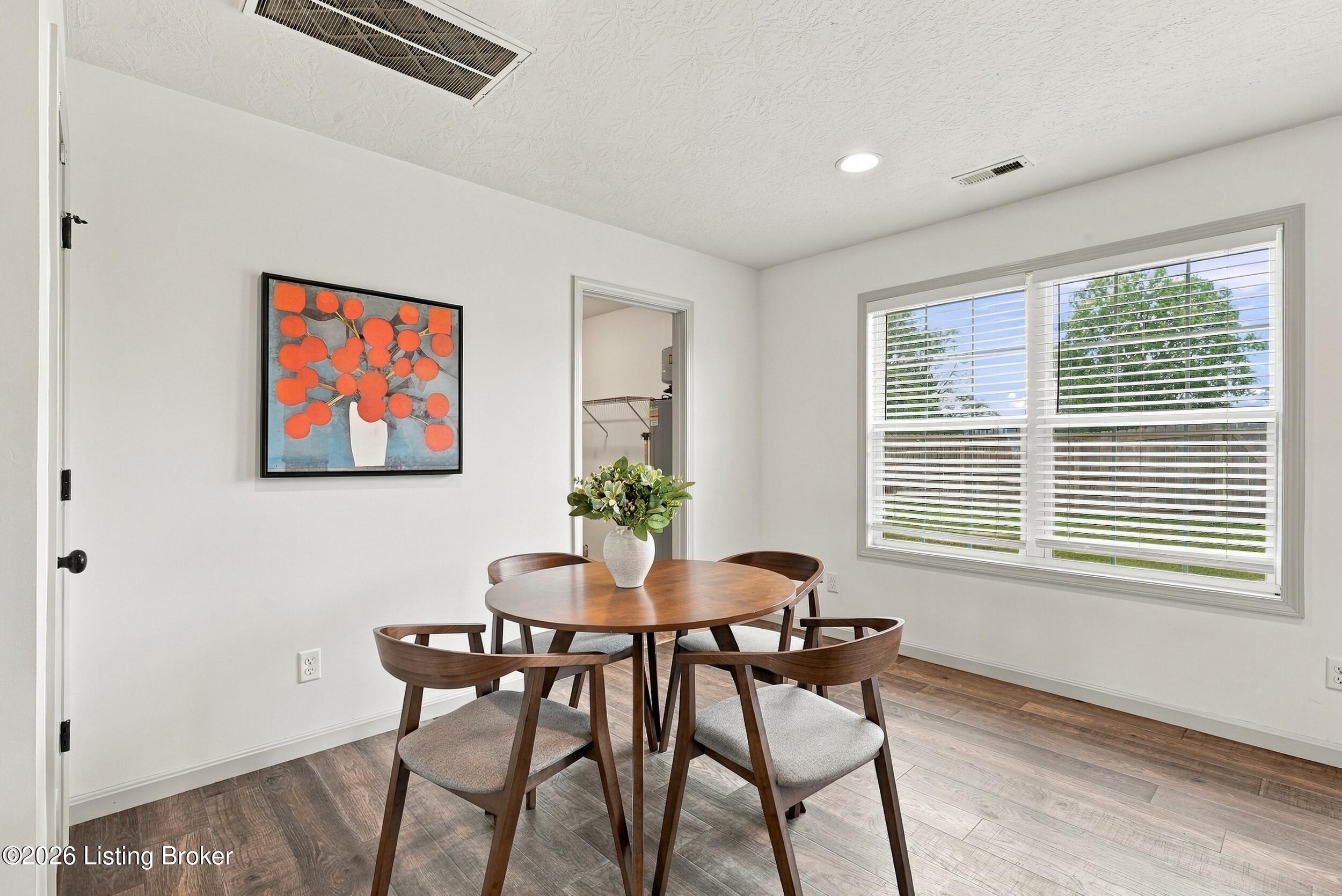 Dining area with a round wooden table, four chairs, colorful wall art, and a large window with blinds.