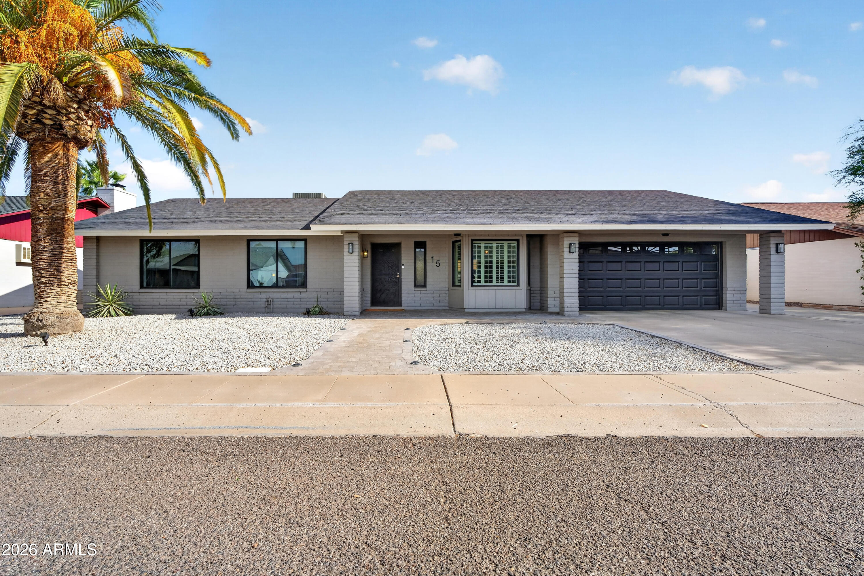 Single-story gray brick home with a dark garage, gravel landscaping, and a large palm tree.