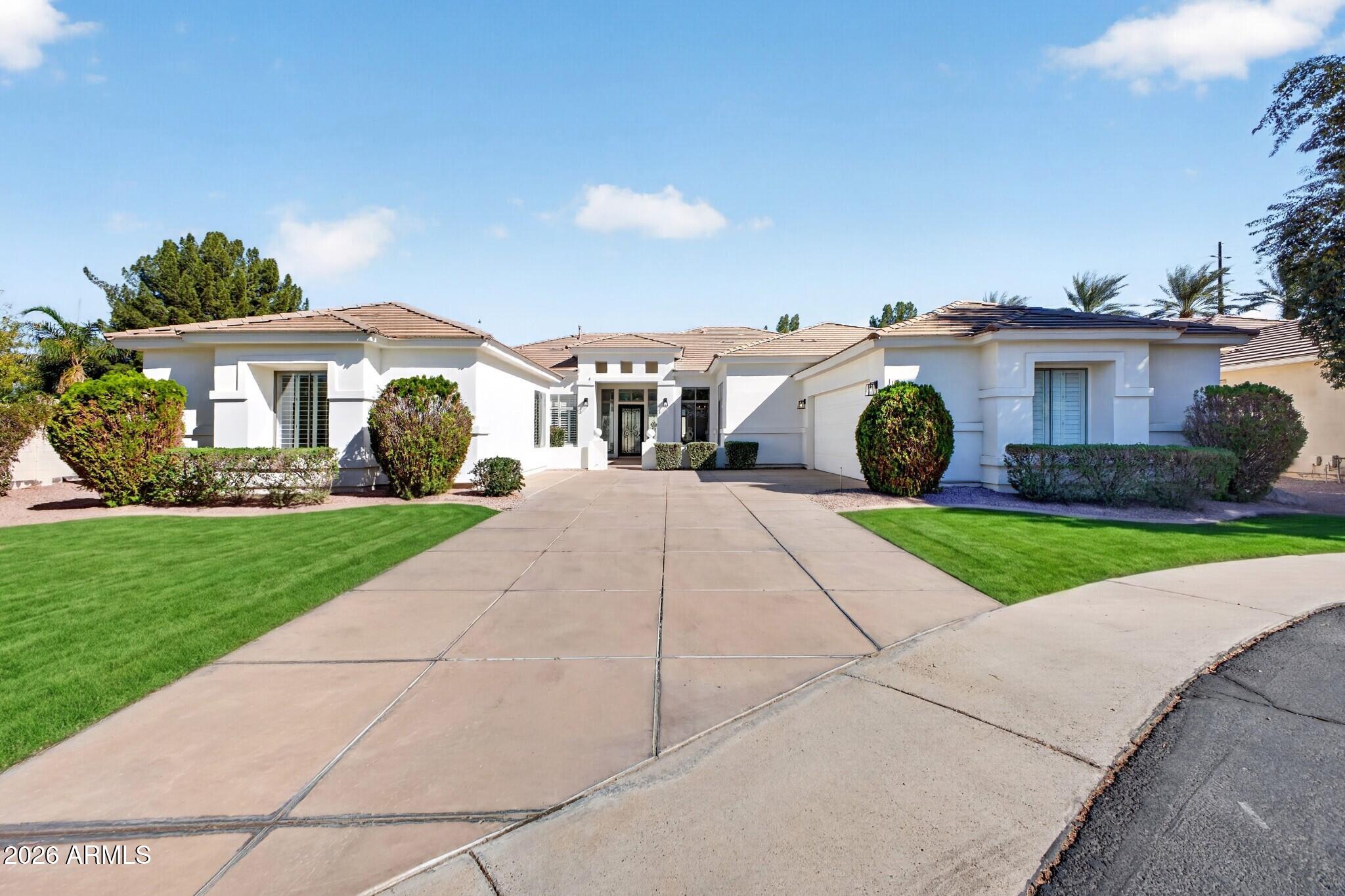 White stucco home with a tile roof, wide driveway, green lawn, and trimmed shrubs.