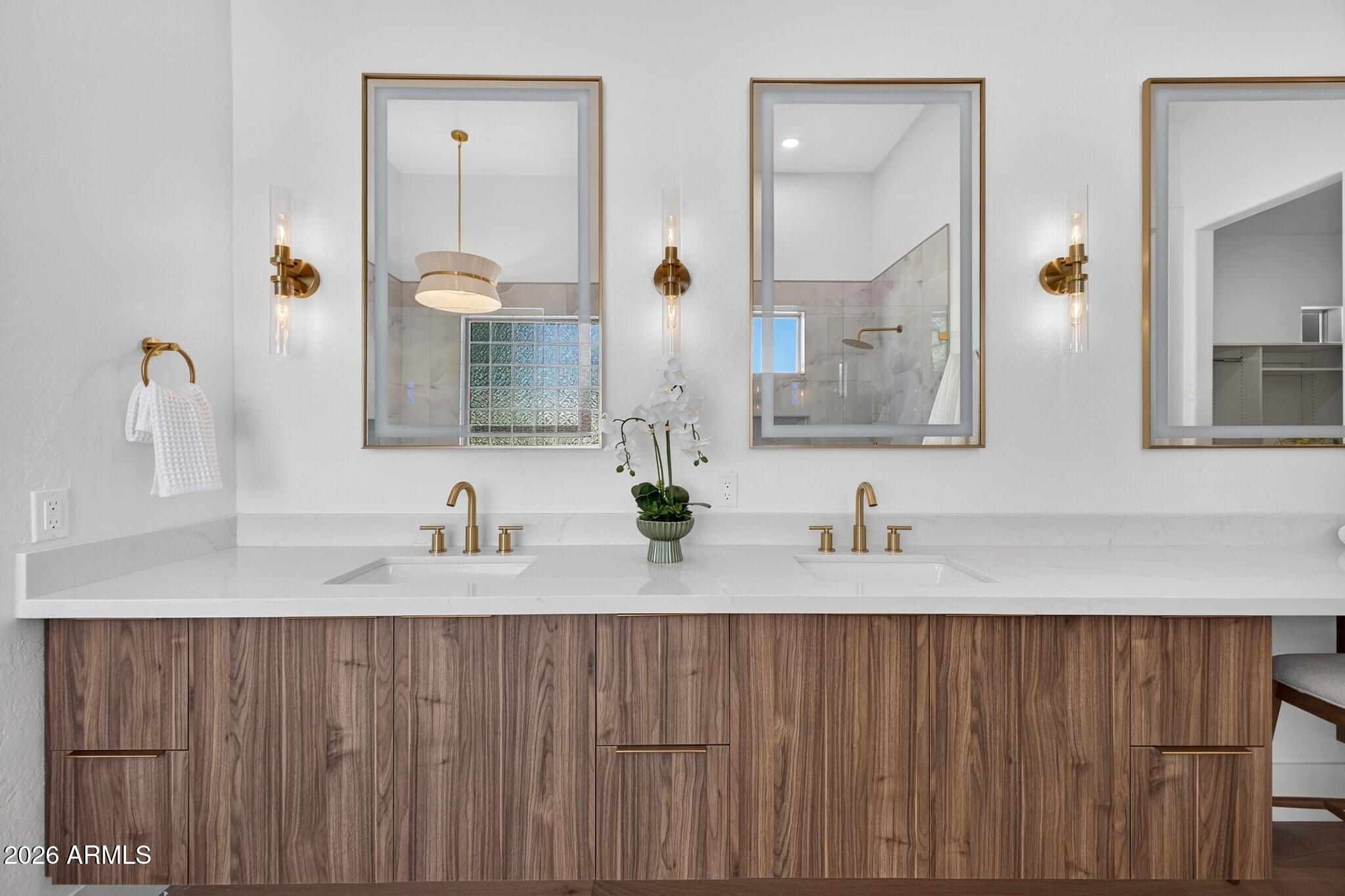 Bathroom double vanity with walnut cabinets, brass fixtures, large mirrors, and a tiled shower reflected behind it.