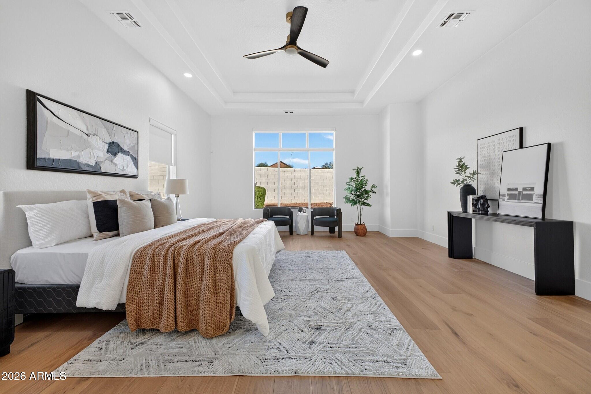 Staged bedroom with a king-size bed, rust-colored throw, tray ceiling, and chairs beside a large window.