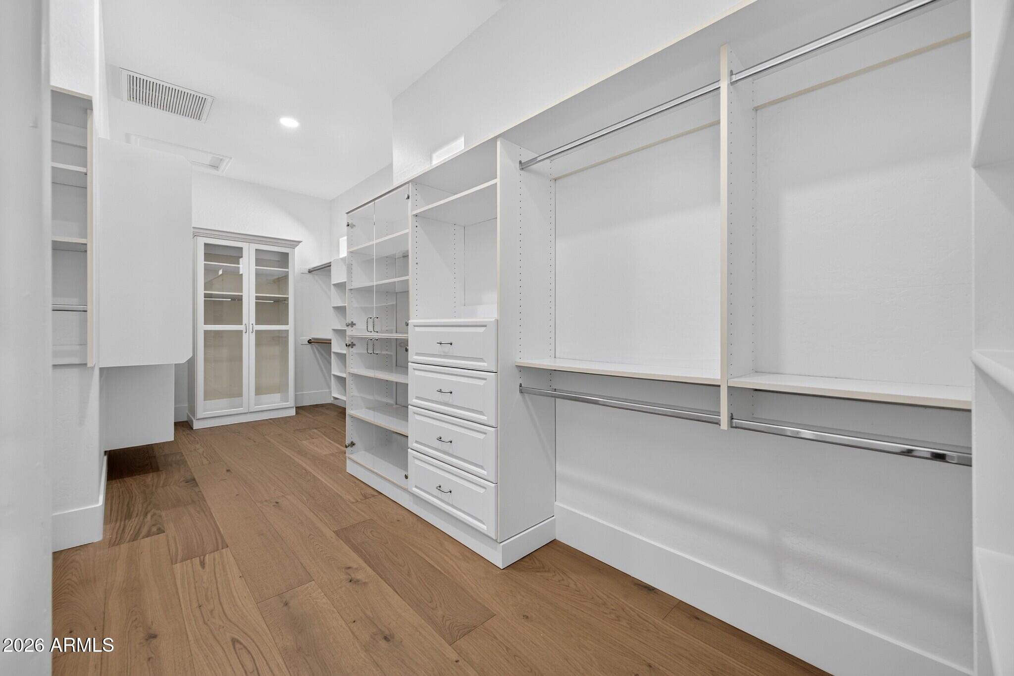 Walk-in closet with white built-in shelving, drawers, a glass-front cabinet, and wood flooring.