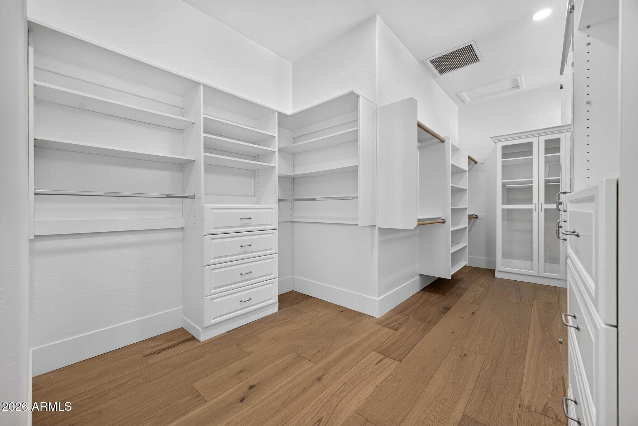Walk-in closet with white built-in shelves, drawers, hanging rails, glass-front cabinets, and wood flooring.