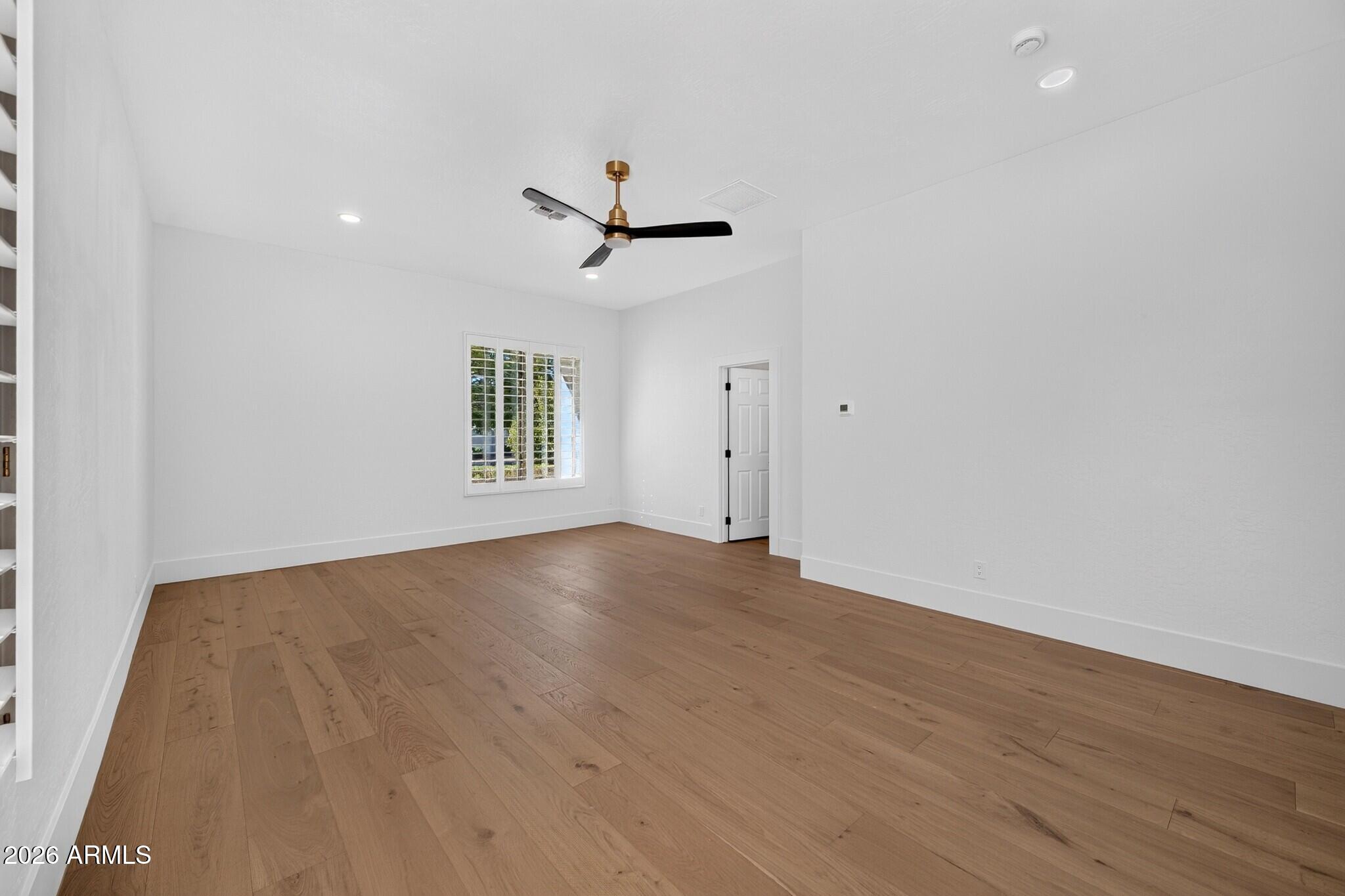 Empty room with wide-plank wood floors, plantation shutters, and a black-and-brass ceiling fan.