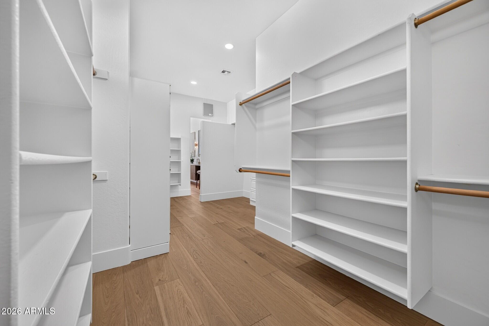 Empty walk-in closet with white built-in shelving, brass-toned hanging rods, and warm wood flooring.