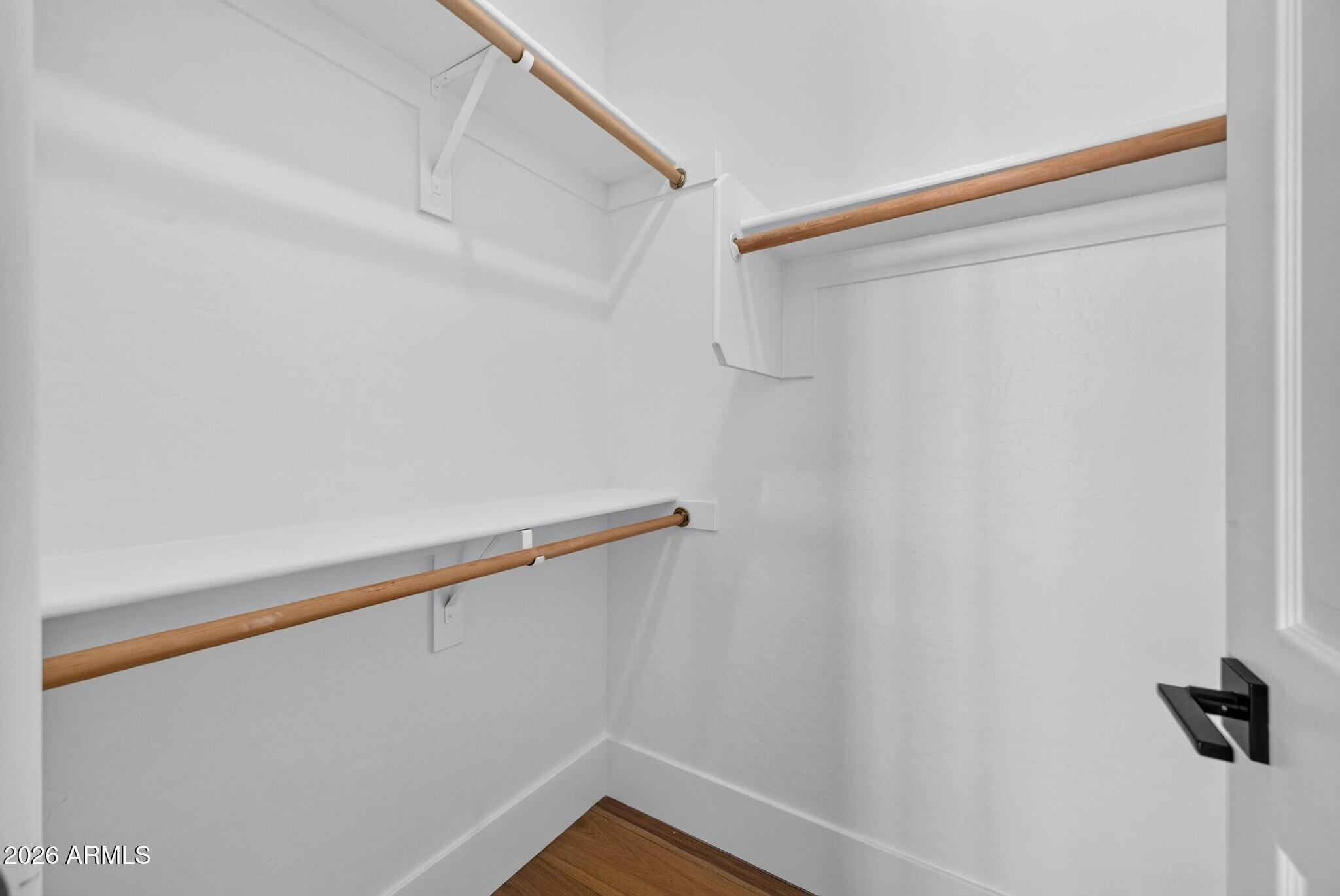 Empty white walk-in closet with wooden hanging rods, a high shelf, and wood flooring.