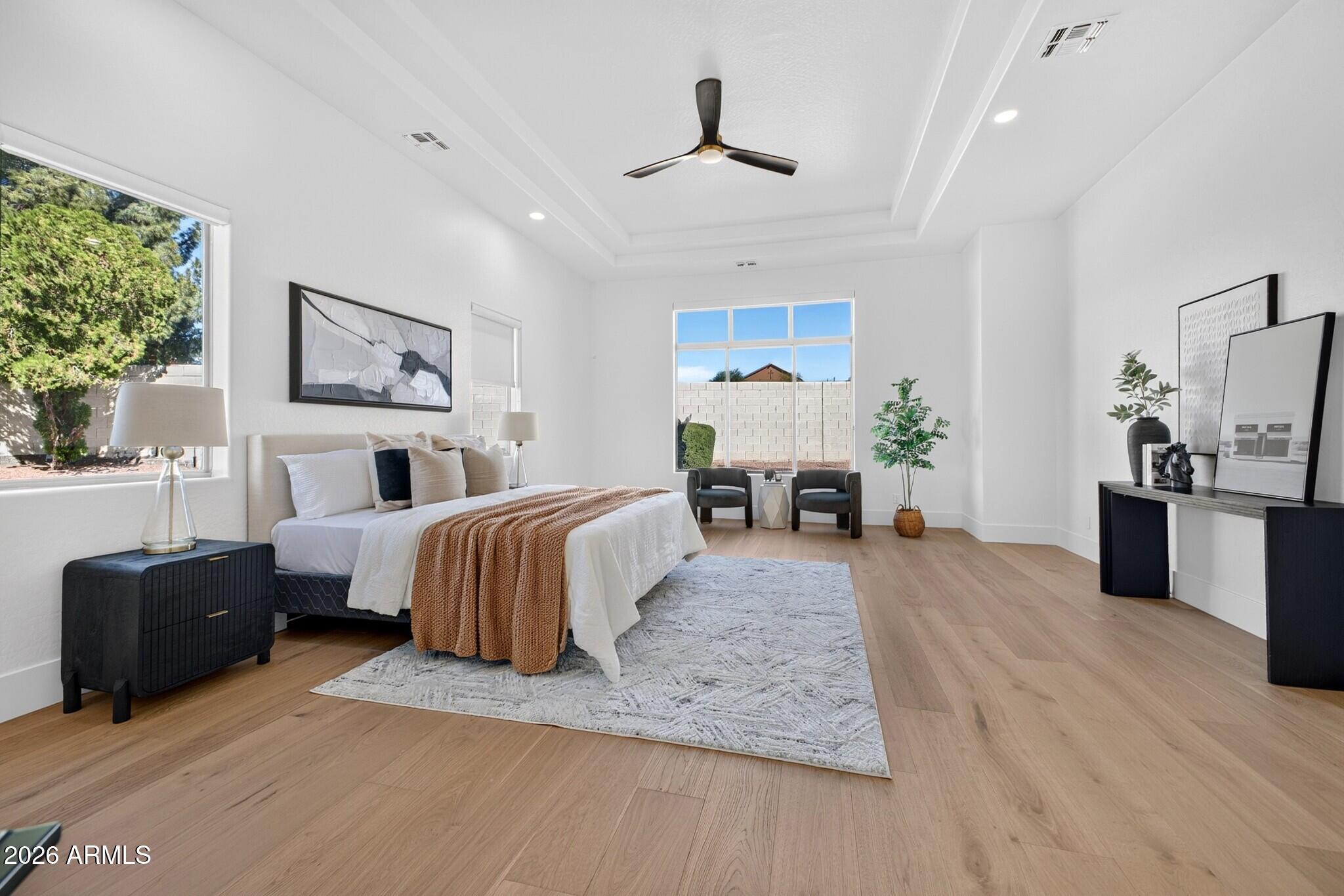 Staged bedroom with a large bed, warm wood floors, tray ceiling, and window-side seating.