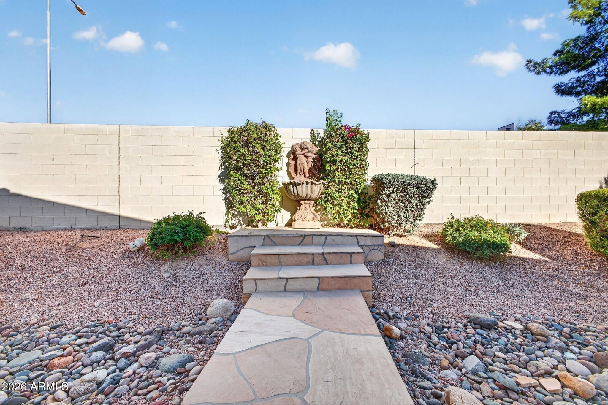 Stone walkway leads to a raised fountain surrounded by gravel, river rocks, and shrubs.