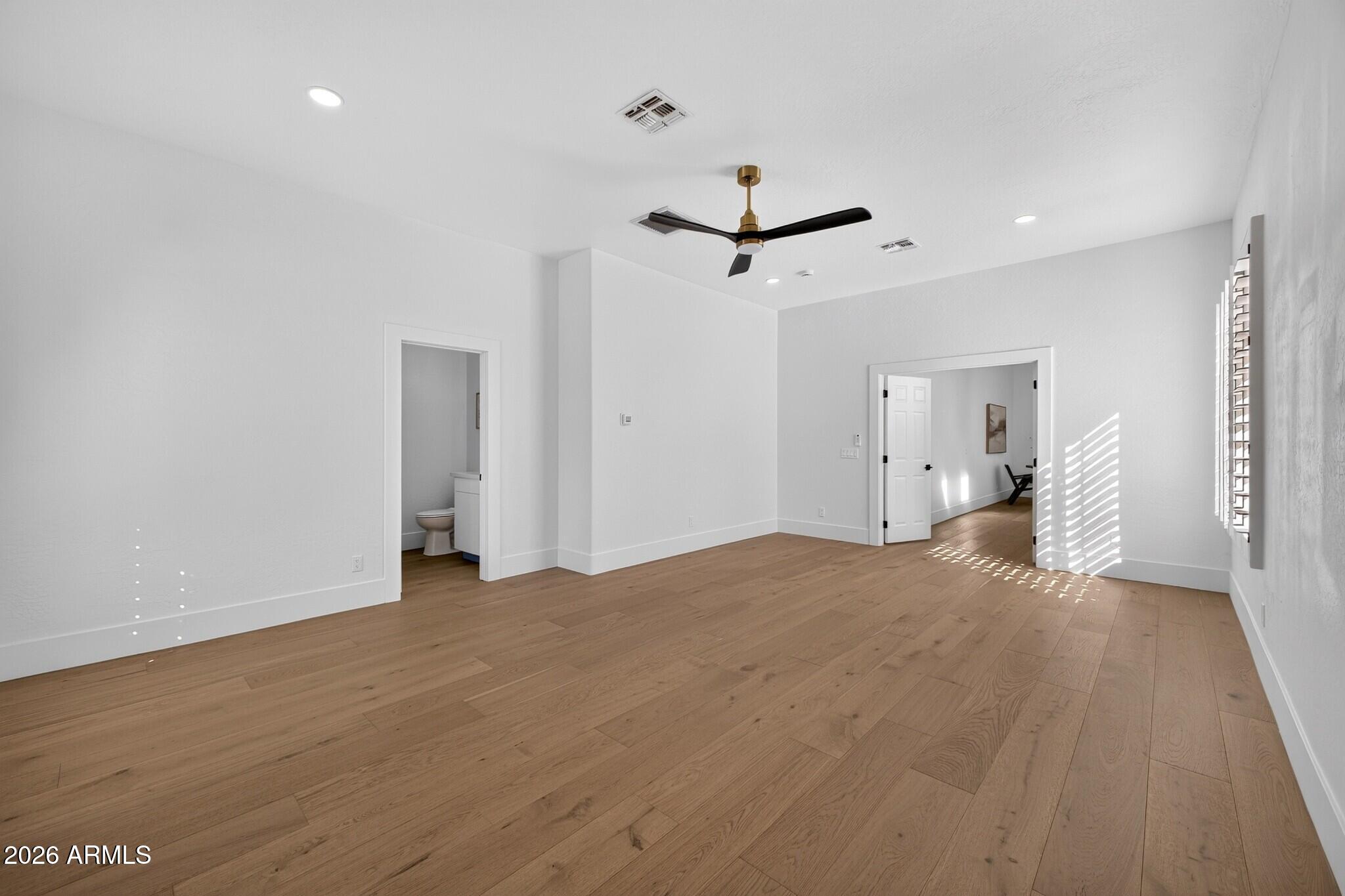 Empty room with wide-plank wood floors, a modern ceiling fan, and sunlight through shutters.
