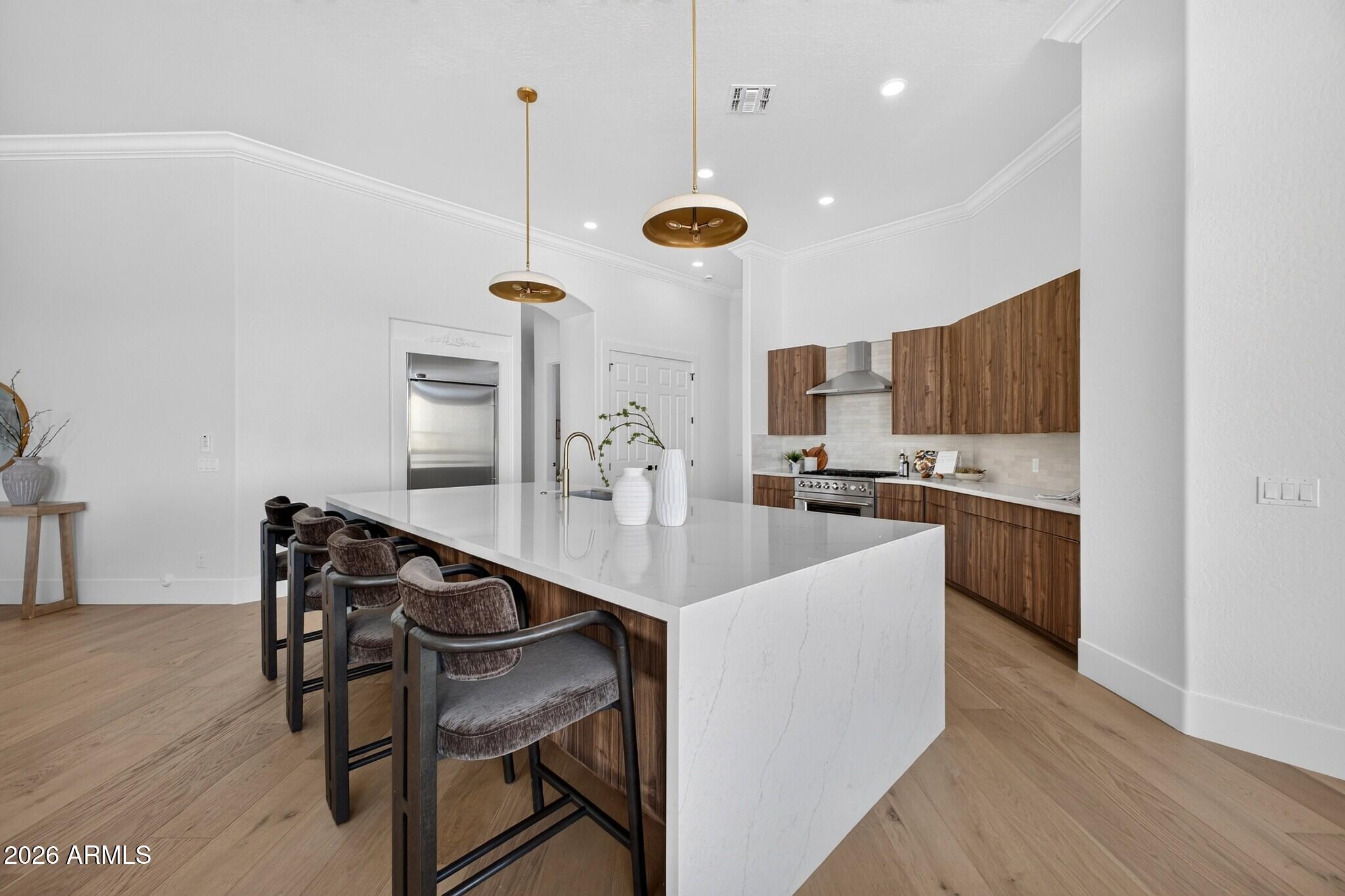 Modern kitchen with a large waterfall island, four upholstered stools, walnut cabinets, and brass pendant lights.