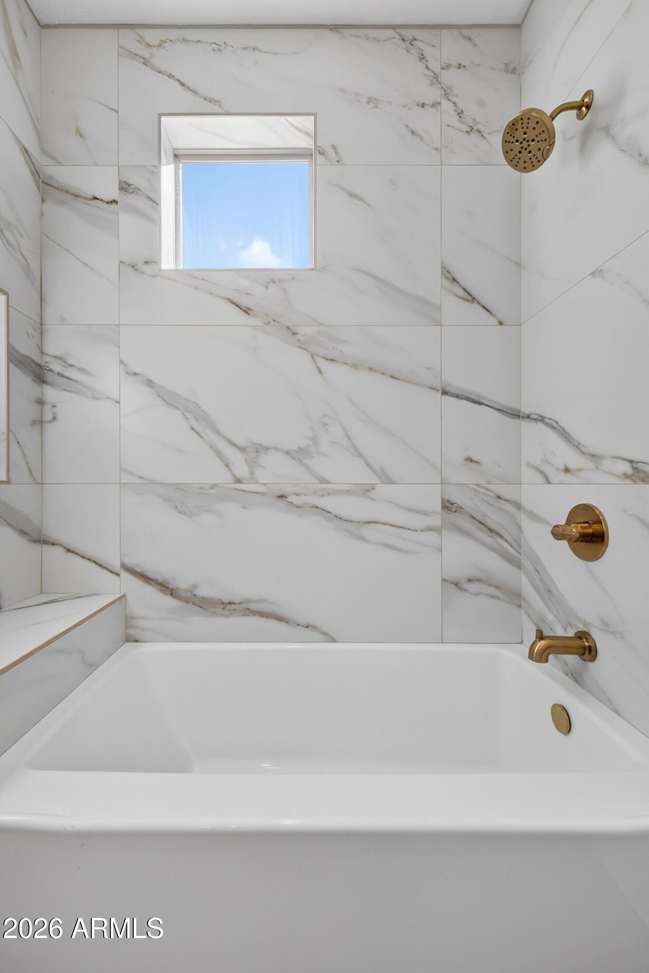 Bathtub with marble-look tile surround, a small window, and brass fixtures.