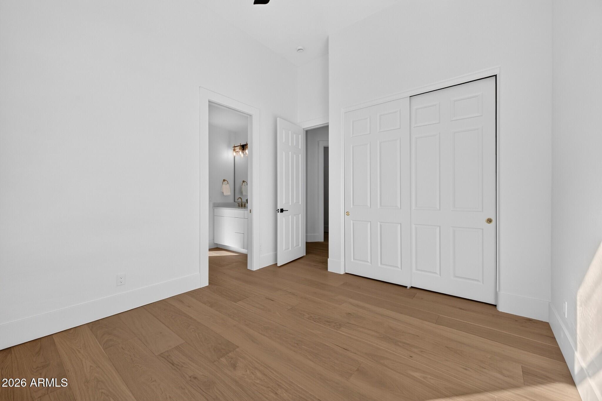 Empty bedroom with wood floors, paneled closet doors, and a bathroom vanity visible through the doorway.