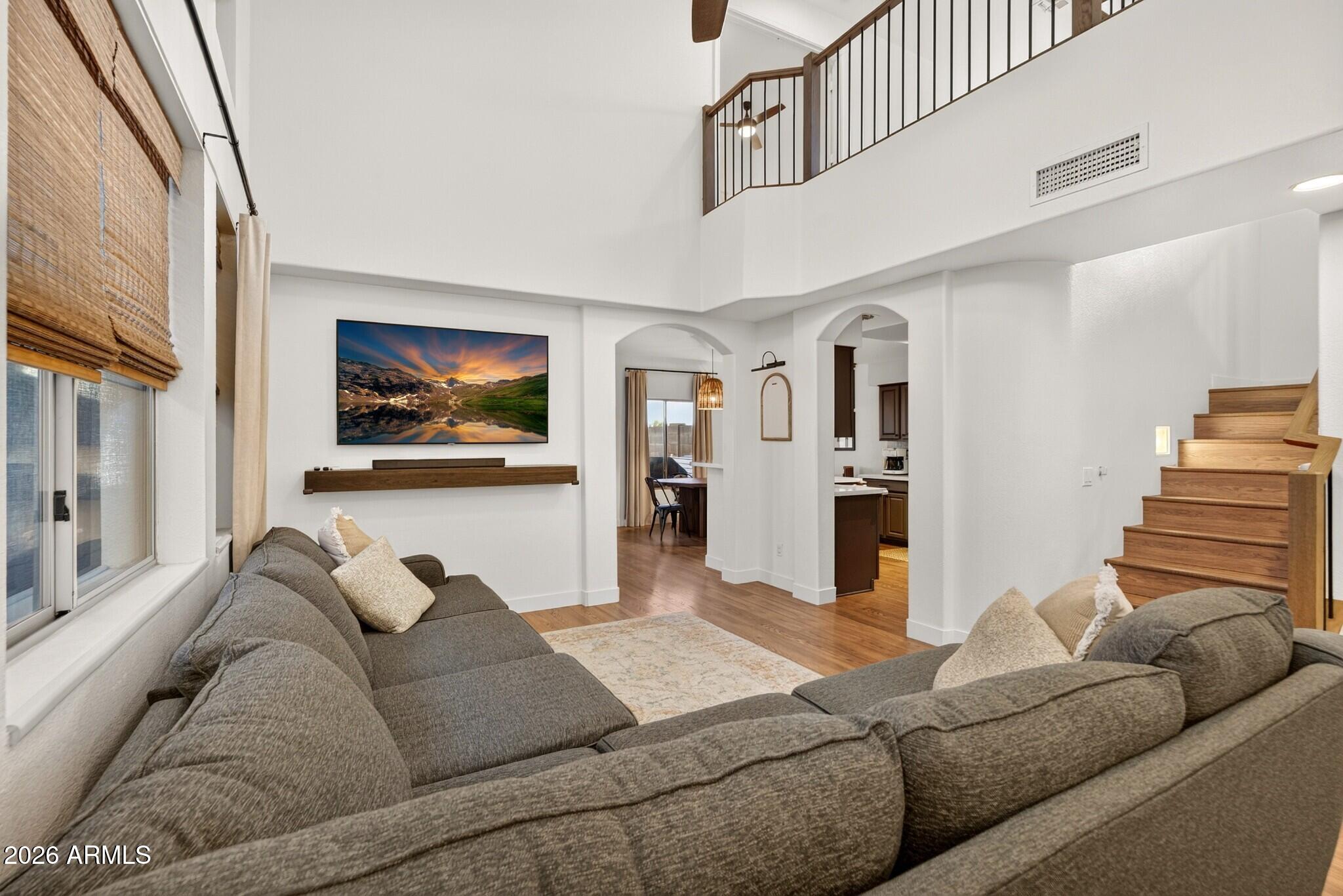 Living room with a tall ceiling, gray sectional sofas, upstairs loft railing, and wooden staircase.