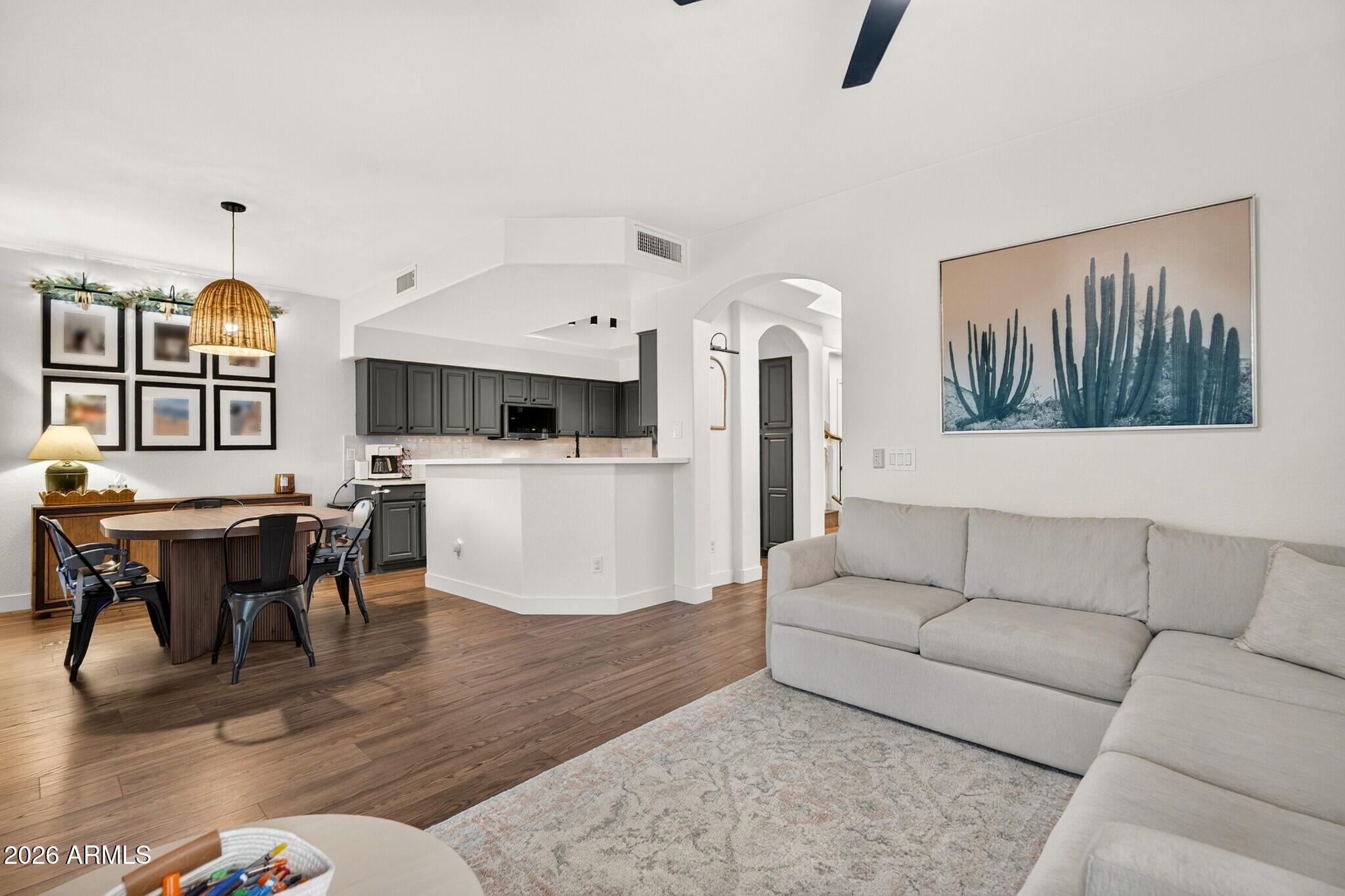 Open-plan living and dining area with a gray sectional, dark kitchen cabinets, and wood floors.