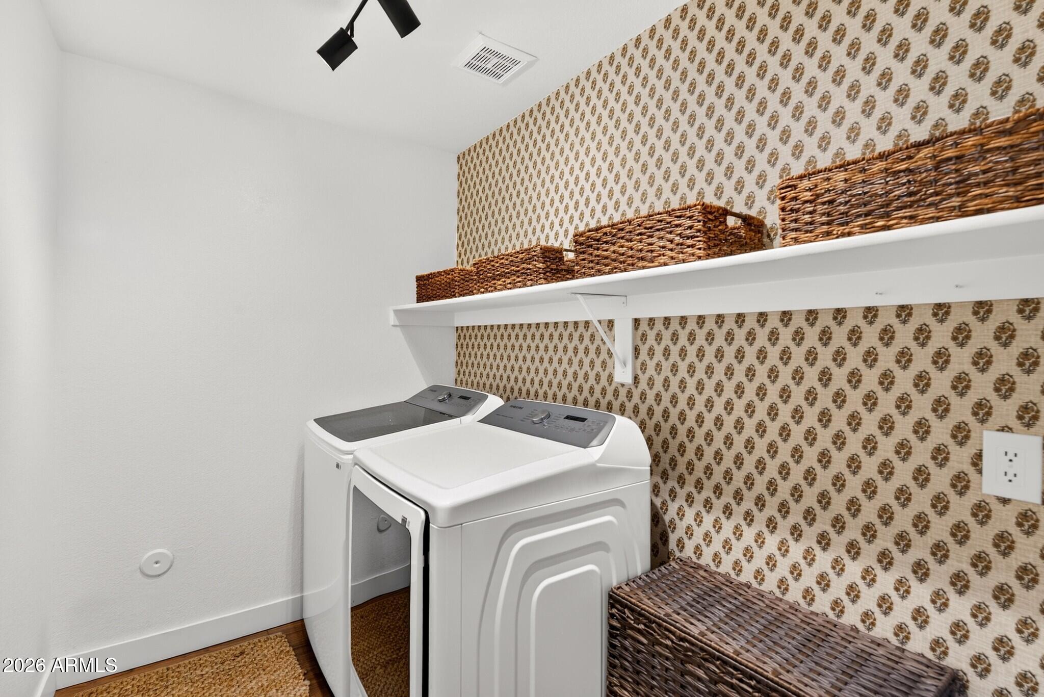 Laundry room with a washer and dryer beneath a shelf of wicker baskets and patterned wallpaper.
