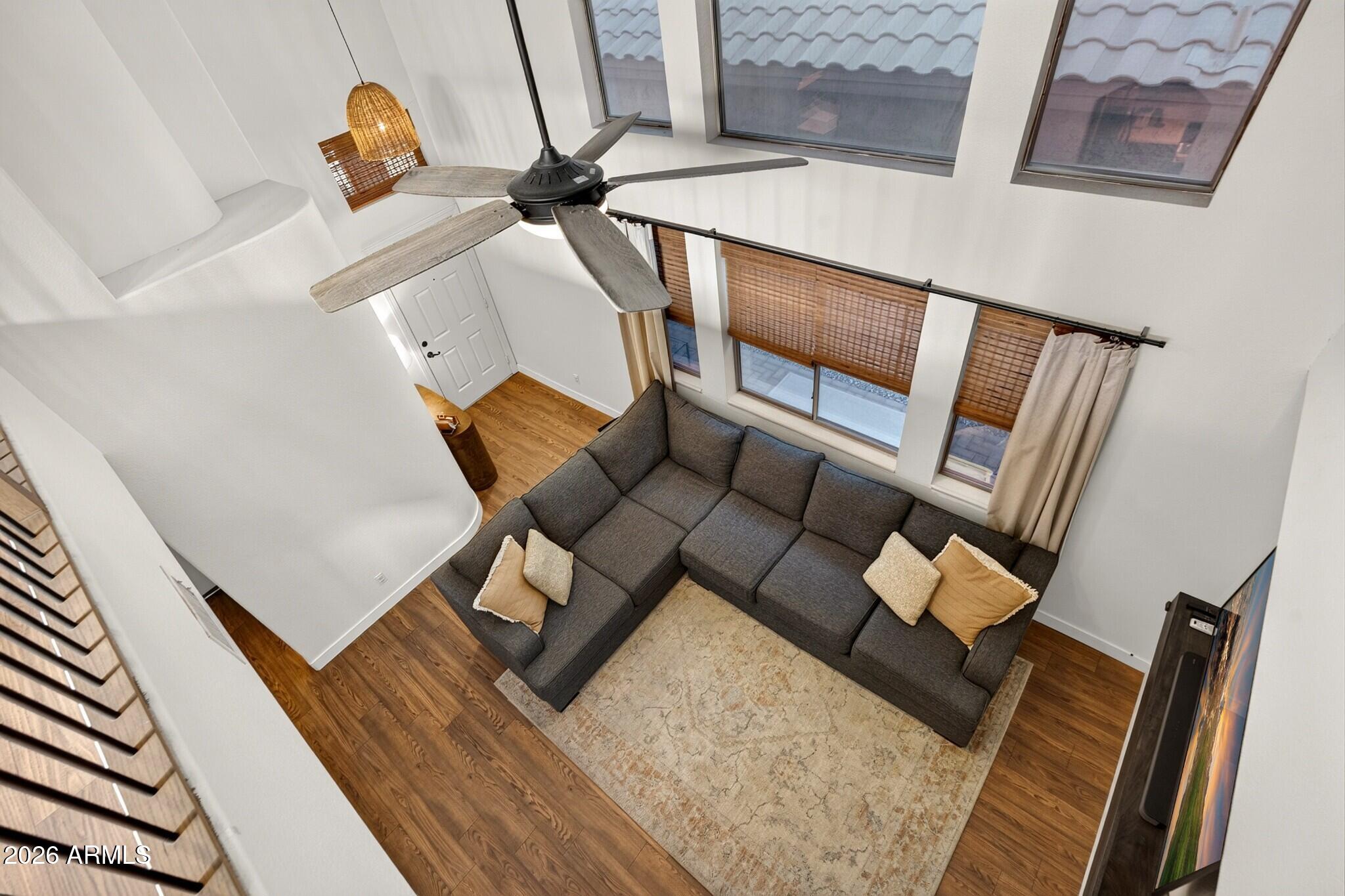 Two-story living room with a gray sectional, tall shaded windows, ceiling fan, and wood flooring.