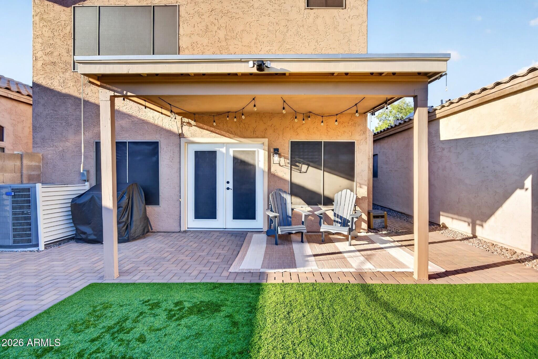 Covered backyard patio with string lights, Adirondack chairs, pavers, and green lawn.