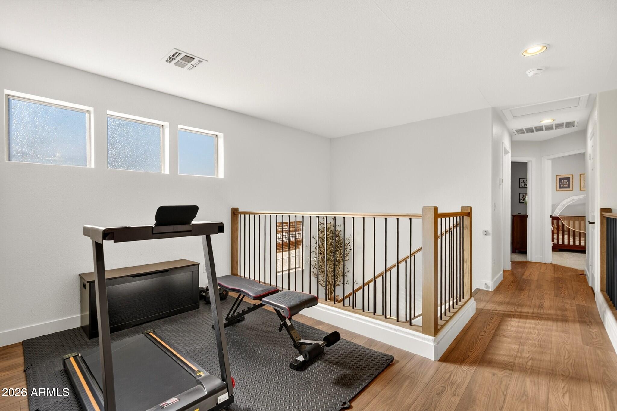 Upstairs home gym with a treadmill and weight bench beside a stair railing and bright high windows.