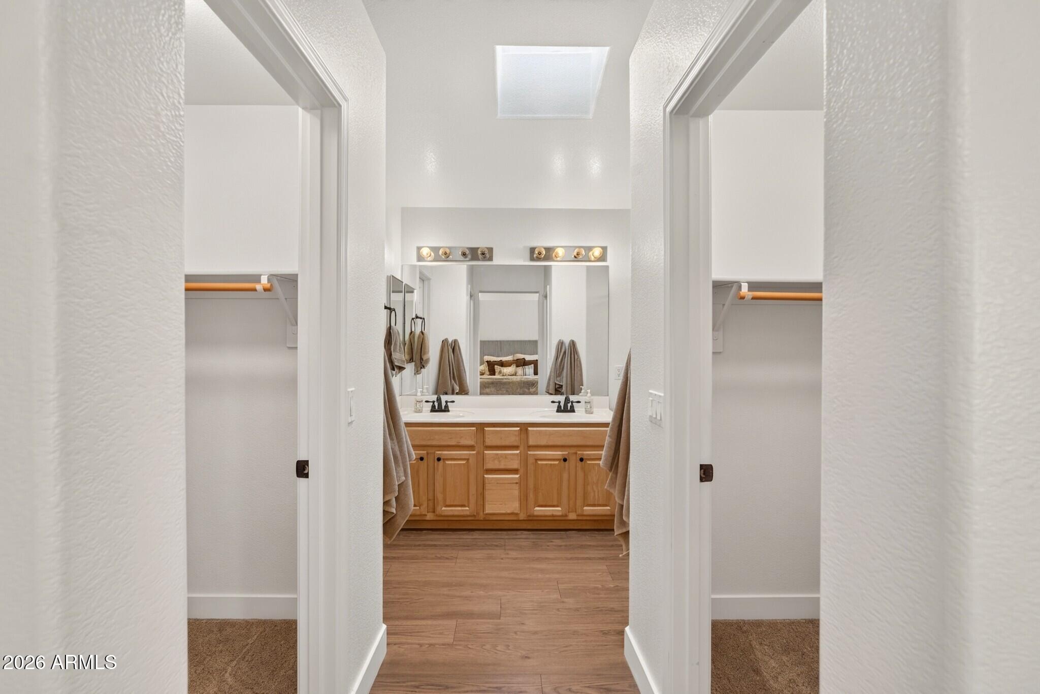 Double-sink bathroom vanity with warm wood cabinets and a skylight, viewed through a hallway.