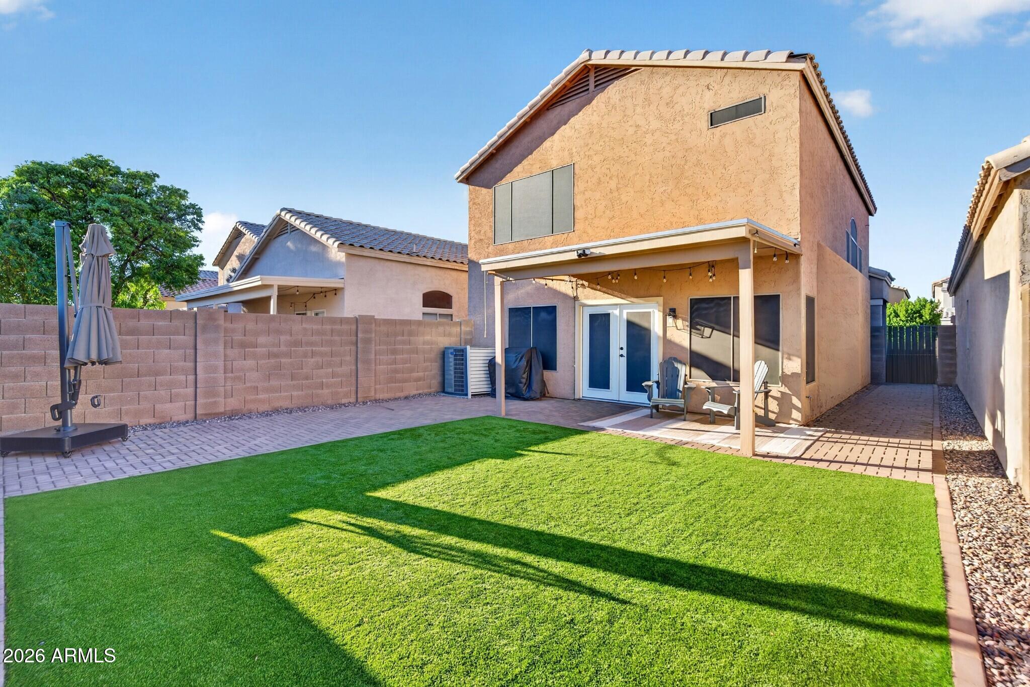 Two-story stucco home with a covered patio, pavers, and a green lawn in the enclosed backyard.